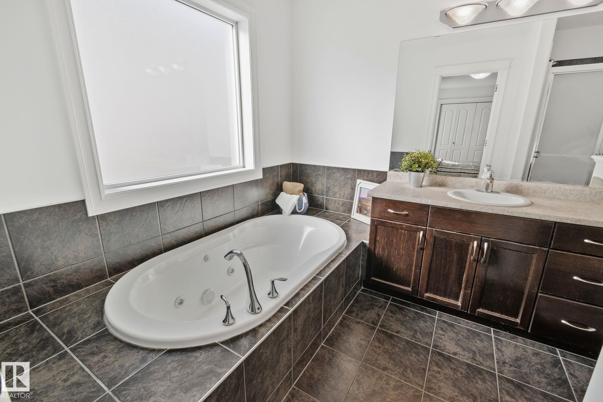 Bathroom featuring a built-in jetted tub, a window with privacy glass, a vanity with a single sink and a mirror, and tiled floors - 1115 61 Street Sw, Edmonton, AB