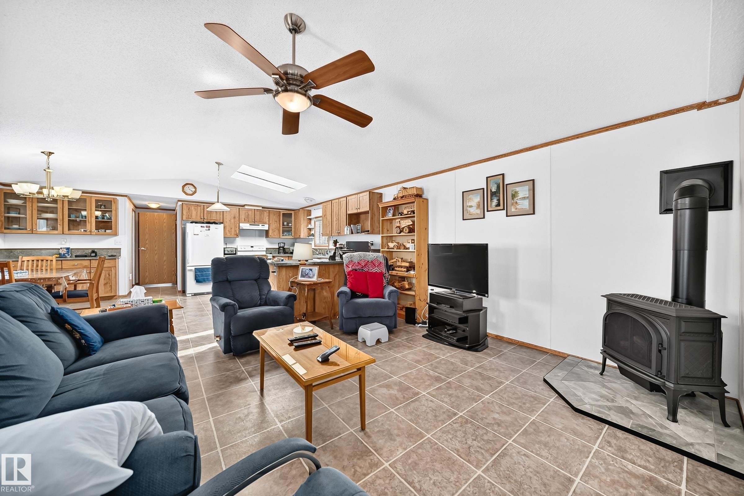 Open-concept living space with tile flooring throughout, featuring a wood-burning stove with a stone hearth, a ceiling fan with integrated lighting, and a kitchen with wood cabinetry and a skylight - #225 3400 48 St, Stony Plain, AB - Indoor Photo Showing Living Room