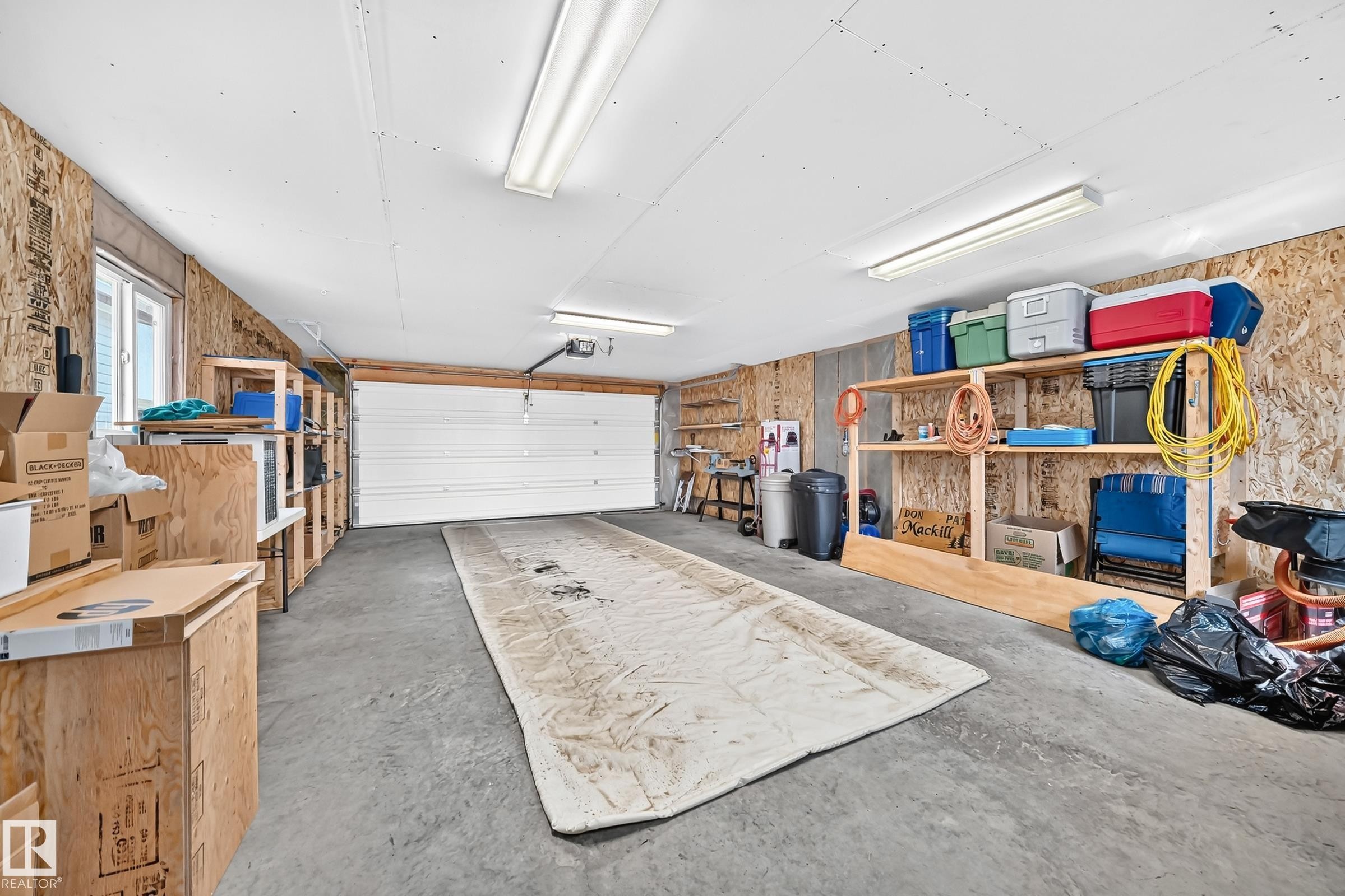 Spacious garage featuring a concrete floor, overhead fluorescent lighting, a white panel garage door, built-in shelving, and a window for natural light - #225 3400 48 St, Stony Plain, AB - Indoor Photo Showing Garage