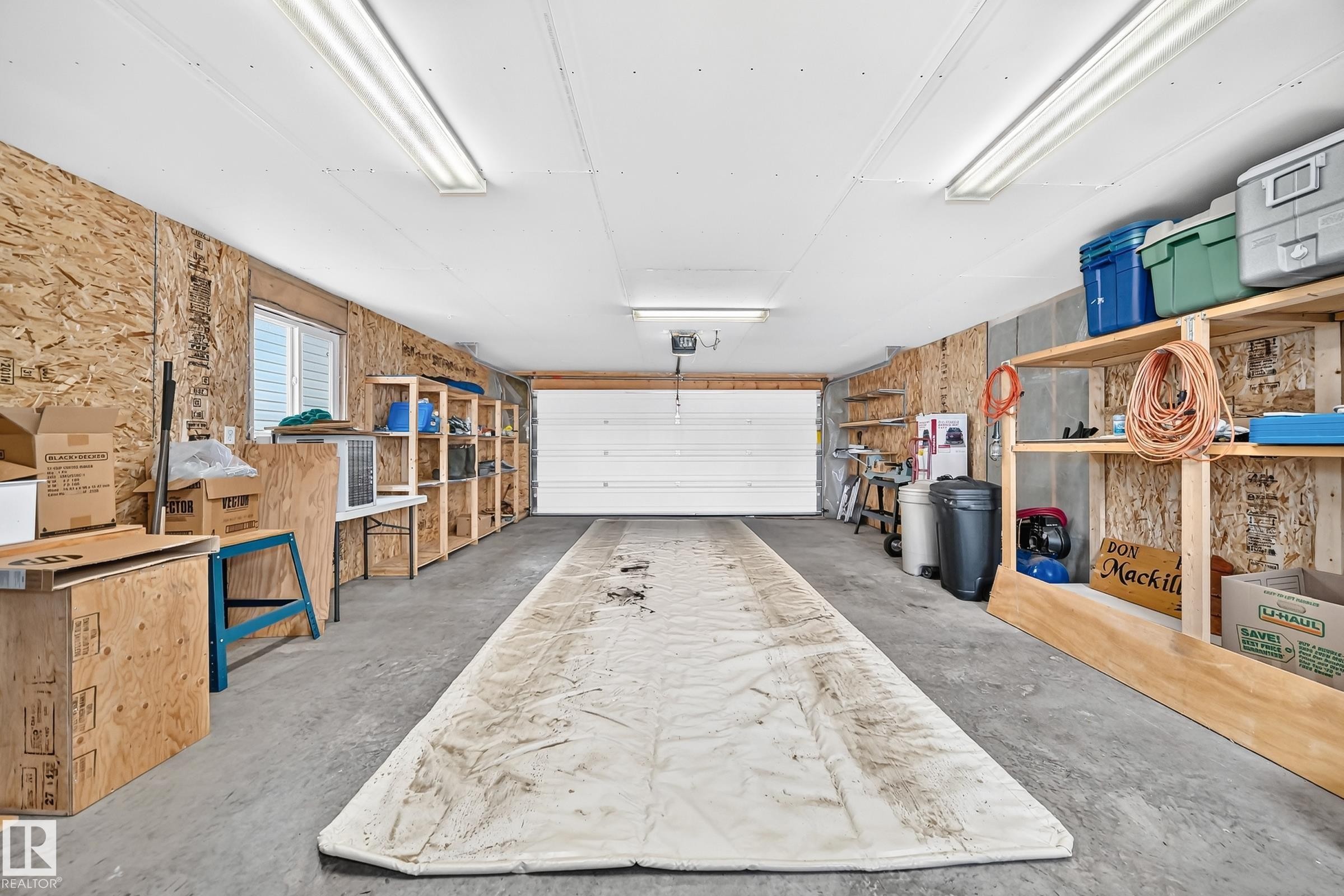 Spacious garage featuring a concrete floor, built-in shelving, white overhead door, and fluorescent lighting - #225 3400 48 St, Stony Plain, AB - Indoor Photo Showing Garage