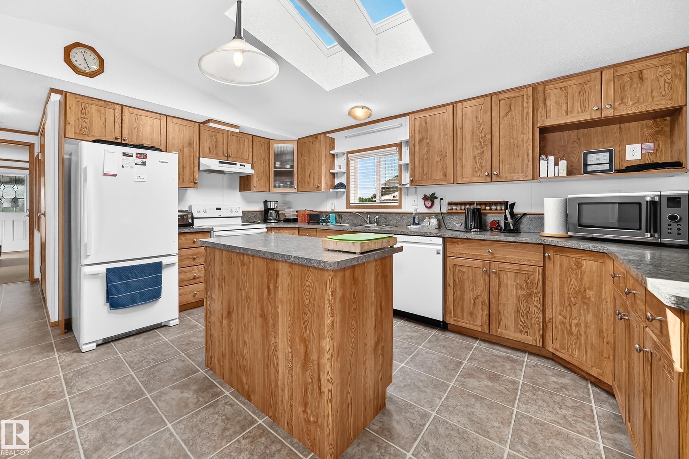 Open kitchen featuring extensive wood-finish cabinetry, a central island with a solid surface countertop, tile flooring, and integrated skylights - #225 3400 48 St, Stony Plain, AB - Indoor Photo Showing Kitchen With Double Sink