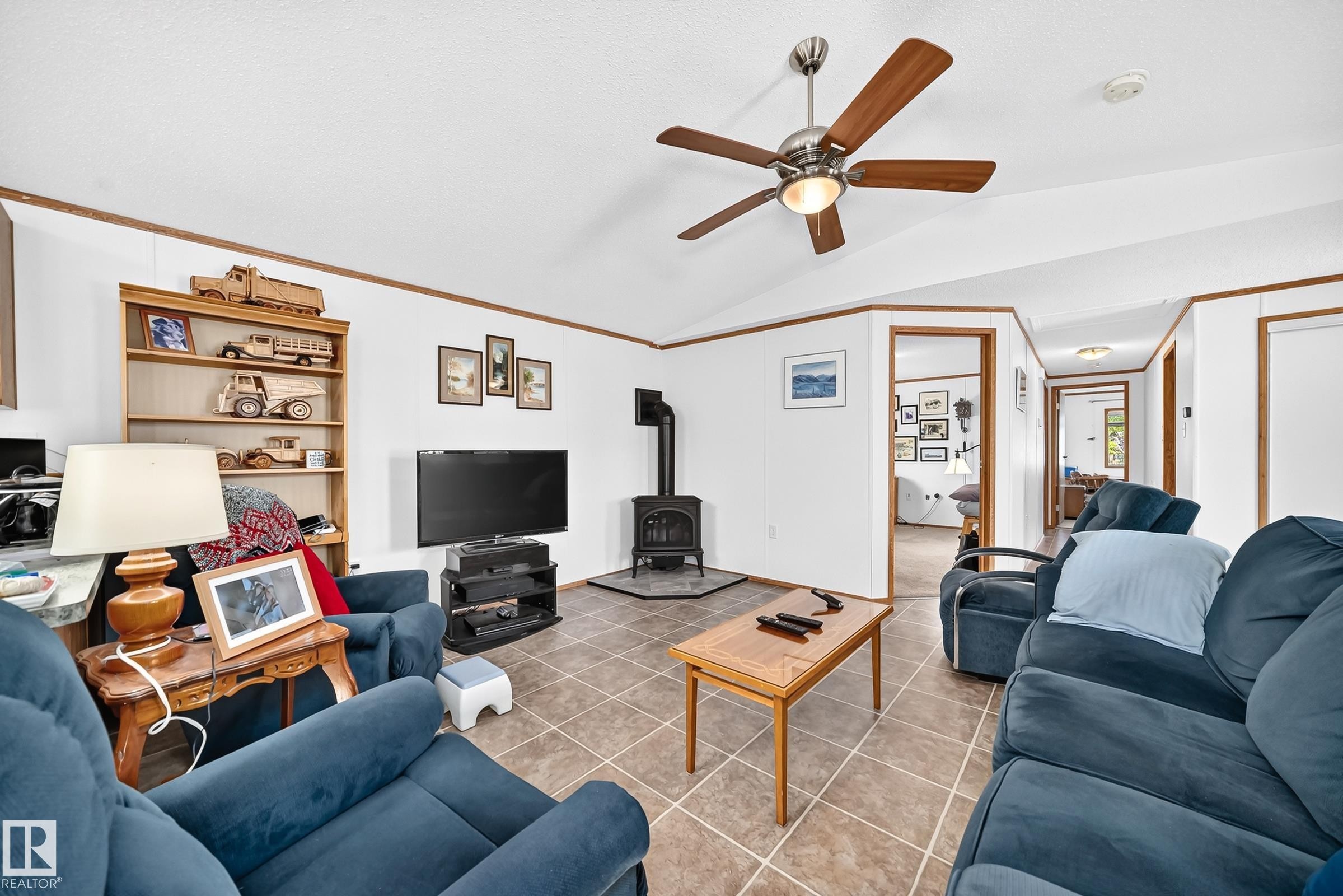 Living area featuring vaulted ceilings, a ceiling fan with light, tiled flooring, a wood-burning stove, and wood-finish trim throughout - #225 3400 48 St, Stony Plain, AB - Indoor Photo Showing Living Room