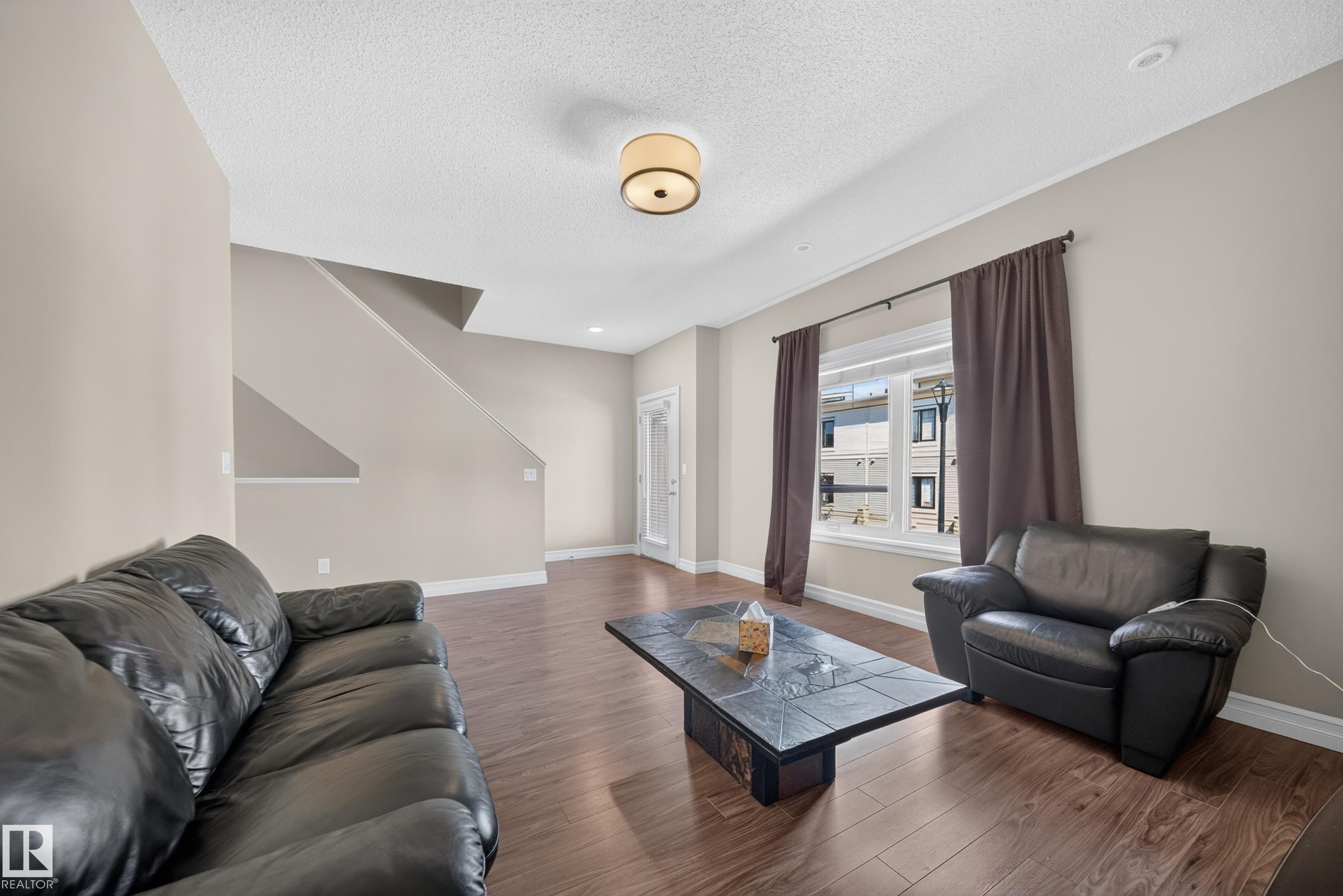 Spacious living area featuring wood-finish flooring, neutral wall tones, a large window, and recessed lighting - #100 804 Welsh Dr Sw, Edmonton, AB - Indoor Photo Showing Living Room