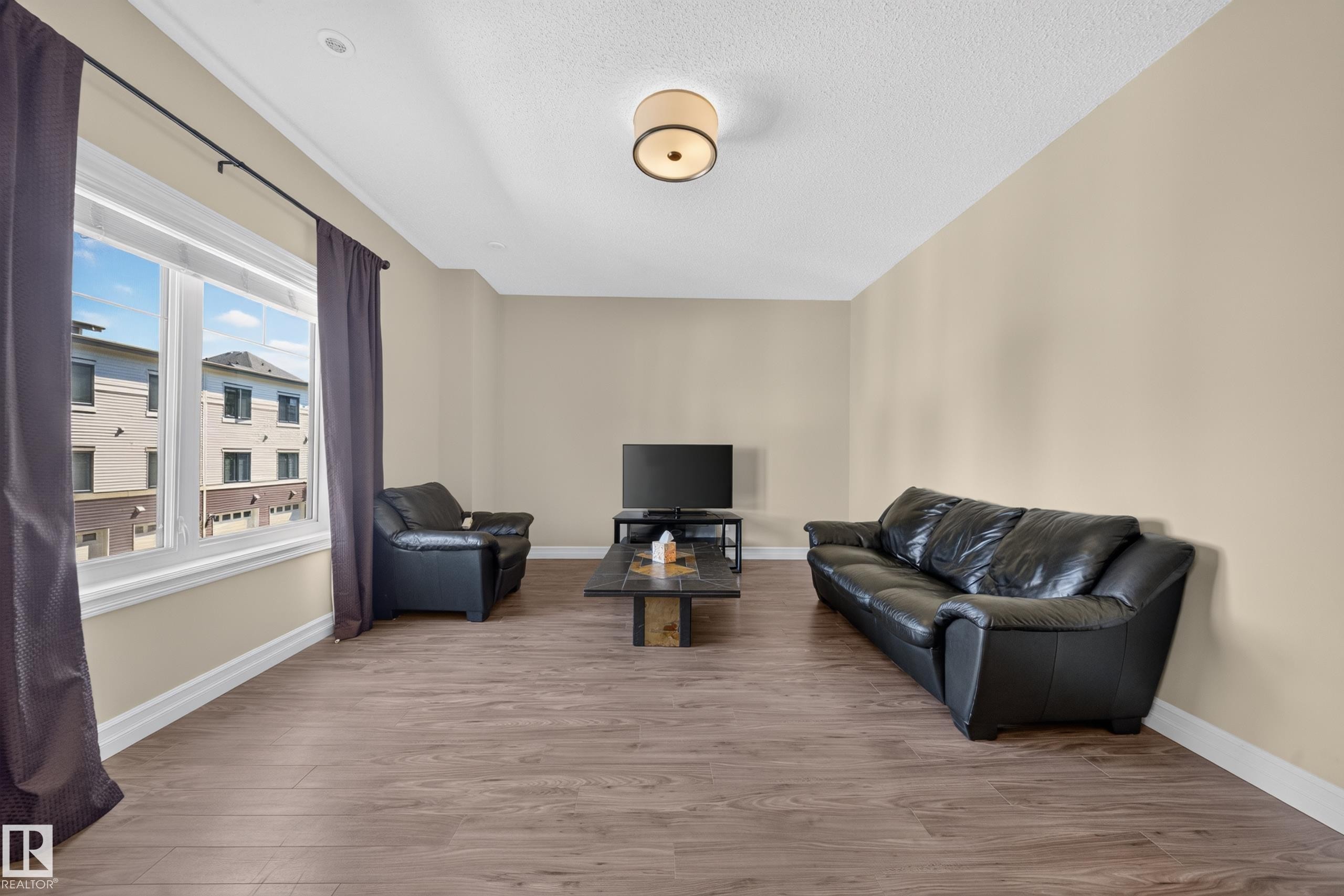 Spacious room featuring wood-finish flooring, neutral-toned walls, and a large window with white trim - #100 804 Welsh Dr Sw, Edmonton, AB - Indoor
