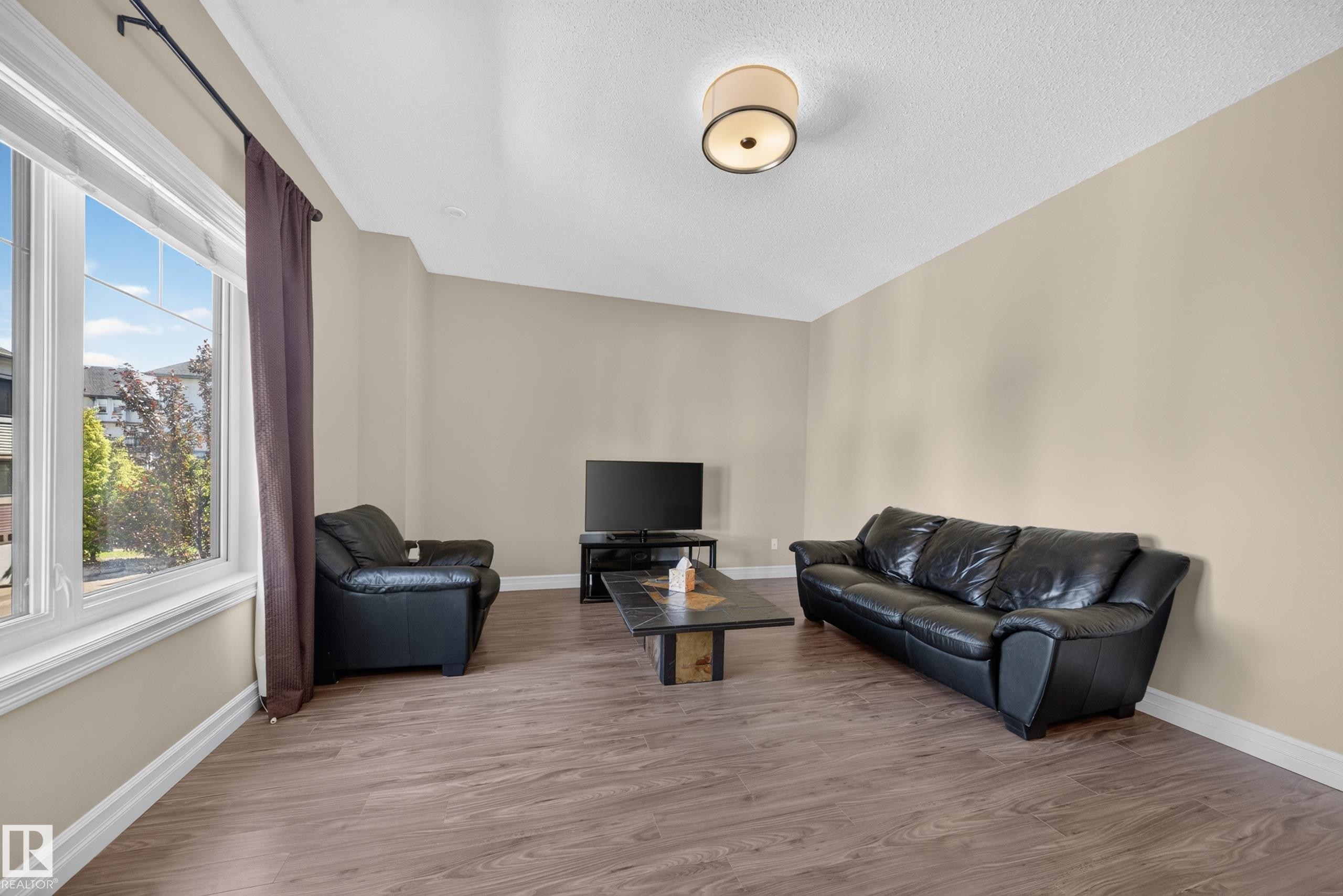 Spacious living area featuring ample natural light from a large window, neutral wall tones, wood-finish flooring, and a flush-mount ceiling light fixture - #100 804 Welsh Dr Sw, Edmonton, AB - Indoor