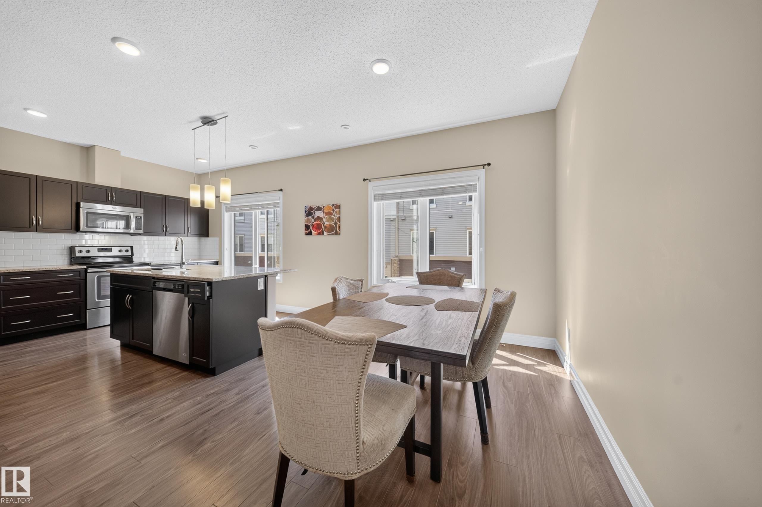 Open-concept kitchen and dining area featuring rich wood-finish flooring, a kitchen island with pendant lighting, stainless steel appliances, and a tiled backsplash - #100 804 Welsh Dr Sw, Edmonton, AB - Indoor