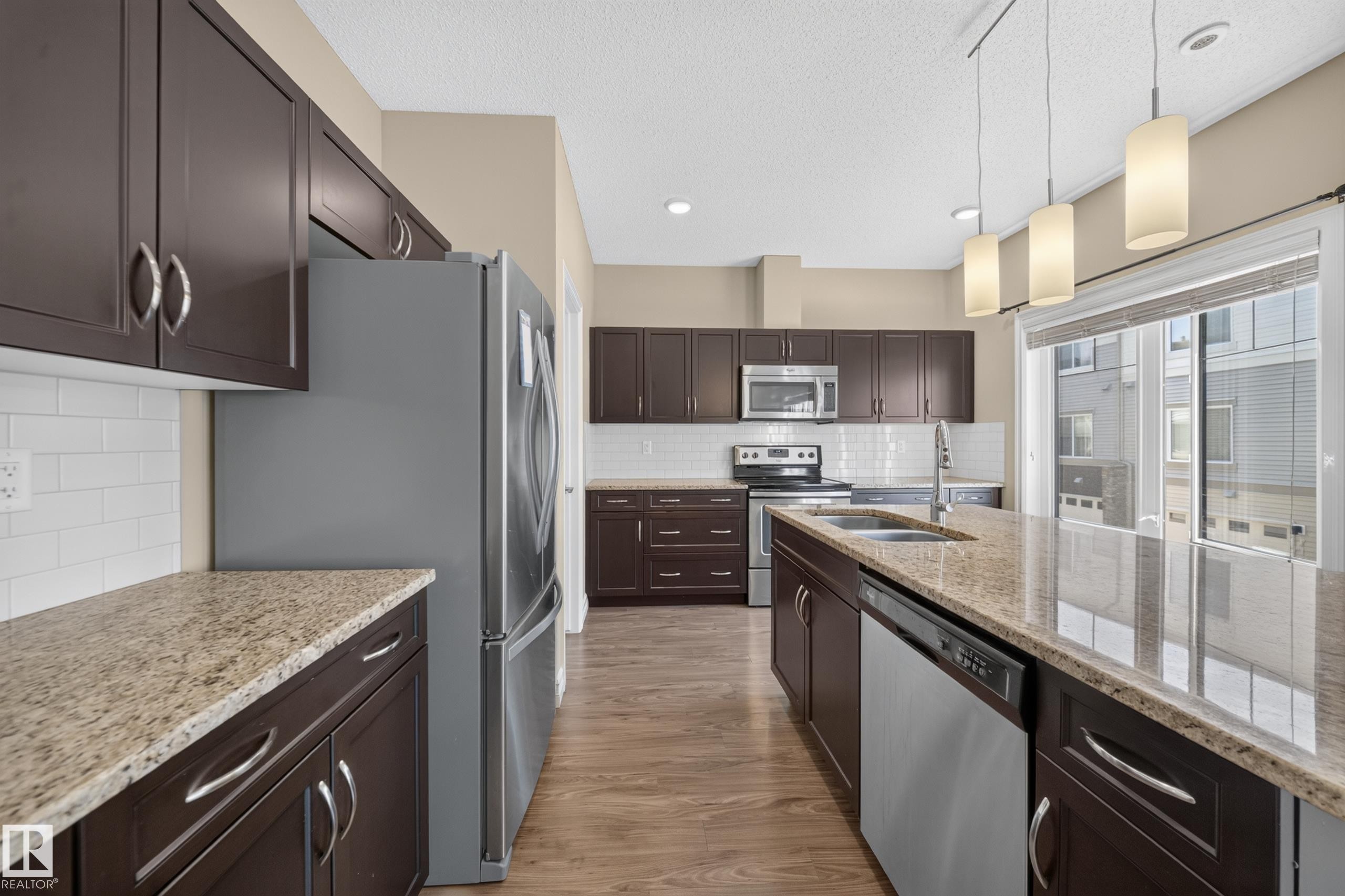 Modern kitchen featuring dark wood cabinetry, granite-finish countertops, stainless steel appliances, a white subway tile backsplash, and wood-finish flooring - #100 804 Welsh Dr Sw, Edmonton, AB - Indoor Photo Showing Kitchen With Stainless Steel Kitchen With Double Sink With Upgraded Kitchen