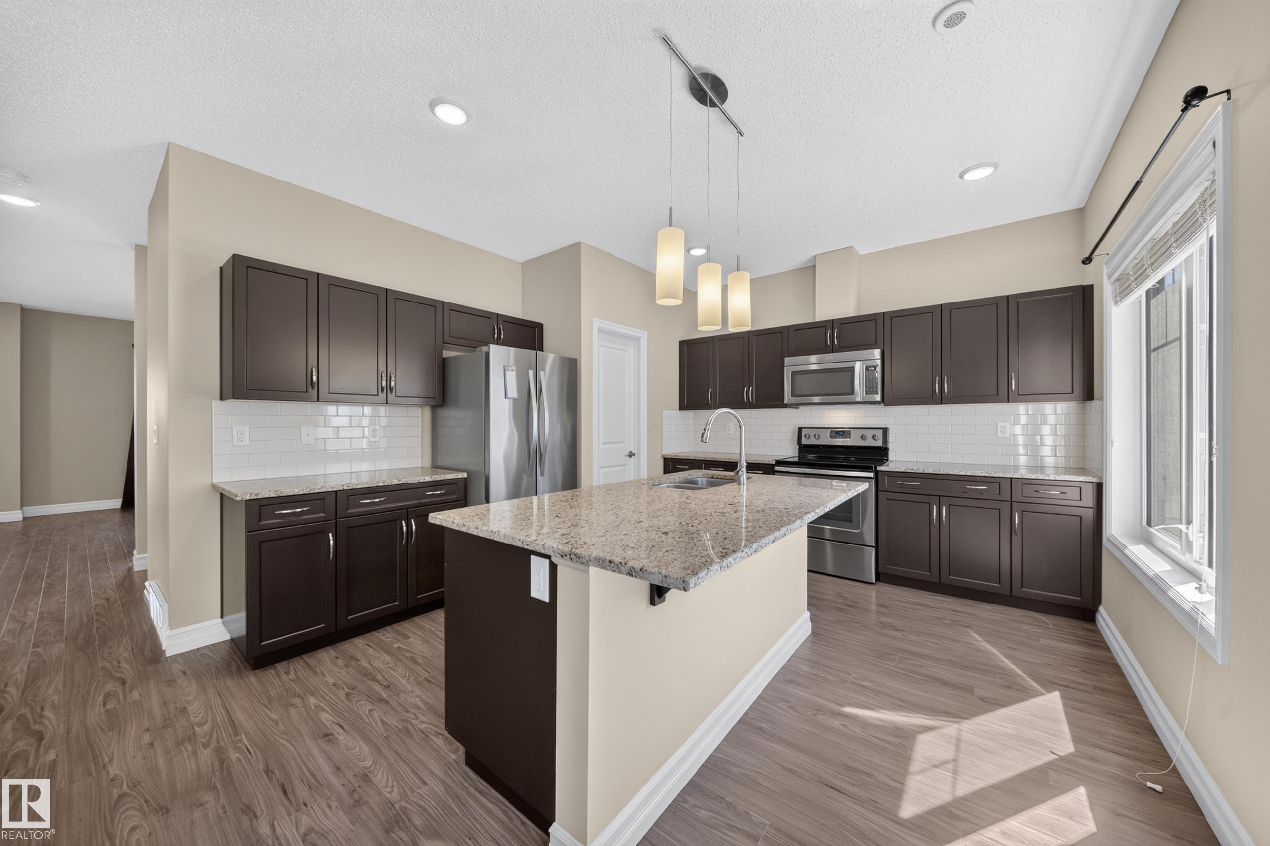 Kitchen featuring wood-finish flooring, dark wood cabinetry with stainless steel hardware, light granite countertops, a white subway tile backsplash, and a central island with pendant lighting - #100 804 Welsh Dr Sw, Edmonton, AB - Indoor Photo Showing Kitchen With Stainless Steel Kitchen With Double Sink With Upgraded Kitchen