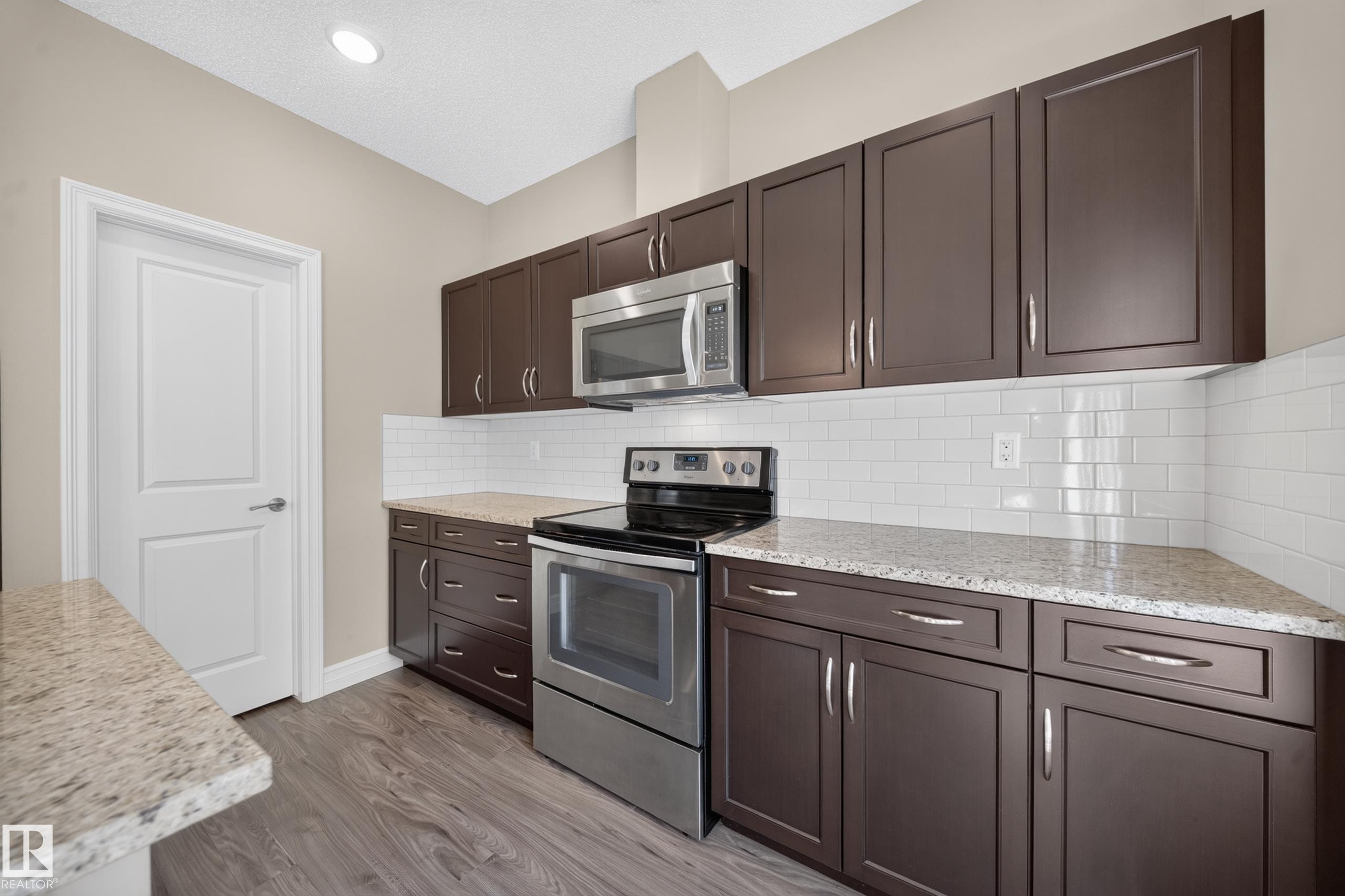 Kitchen featuring dark wood-finish cabinetry with silver-tone hardware, granite-look countertops, and white subway tile backsplash - #100 804 Welsh Dr Sw, Edmonton, AB - Indoor Photo Showing Kitchen