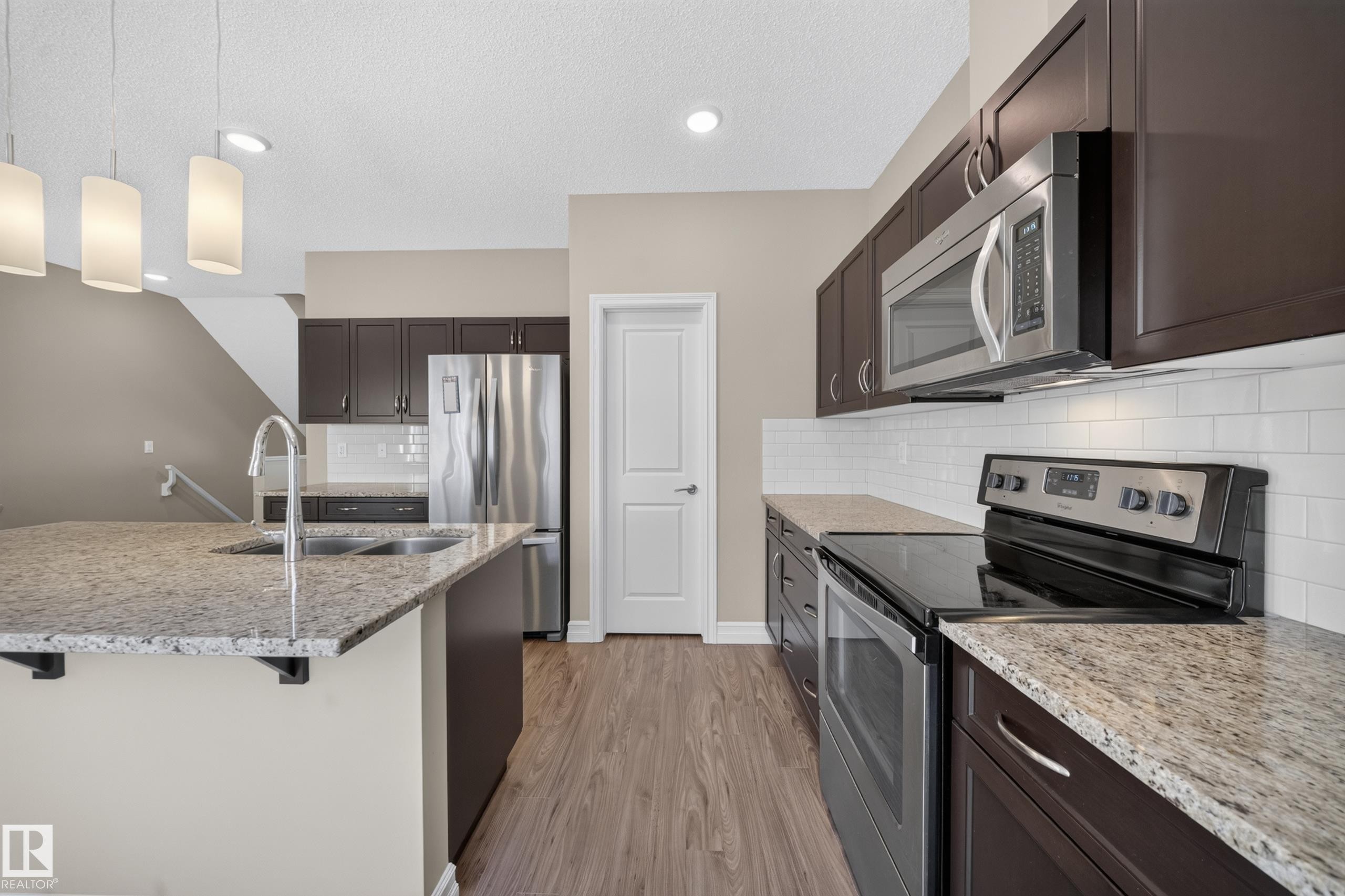 Modern kitchen featuring dark wood cabinetry, light granite countertops, stainless steel appliances, and wood-finish flooring - #100 804 Welsh Dr Sw, Edmonton, AB - Indoor Photo Showing Kitchen With Stainless Steel Kitchen With Double Sink With Upgraded Kitchen