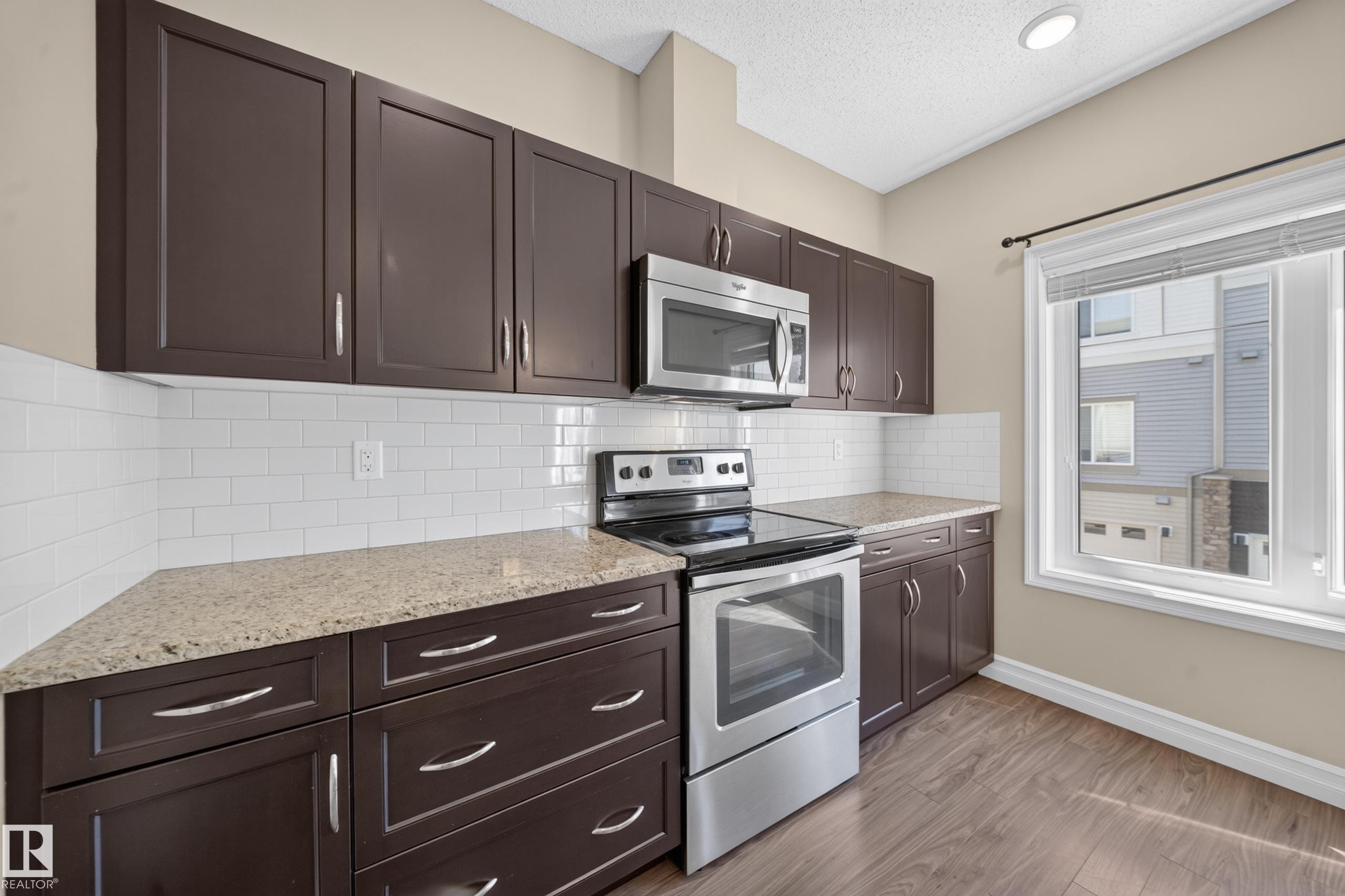 Kitchen featuring dark wood cabinetry, light-toned countertops, white subway tile backsplash, and stainless steel appliances - #100 804 Welsh Dr Sw, Edmonton, AB - Indoor Photo Showing Kitchen With Stainless Steel Kitchen