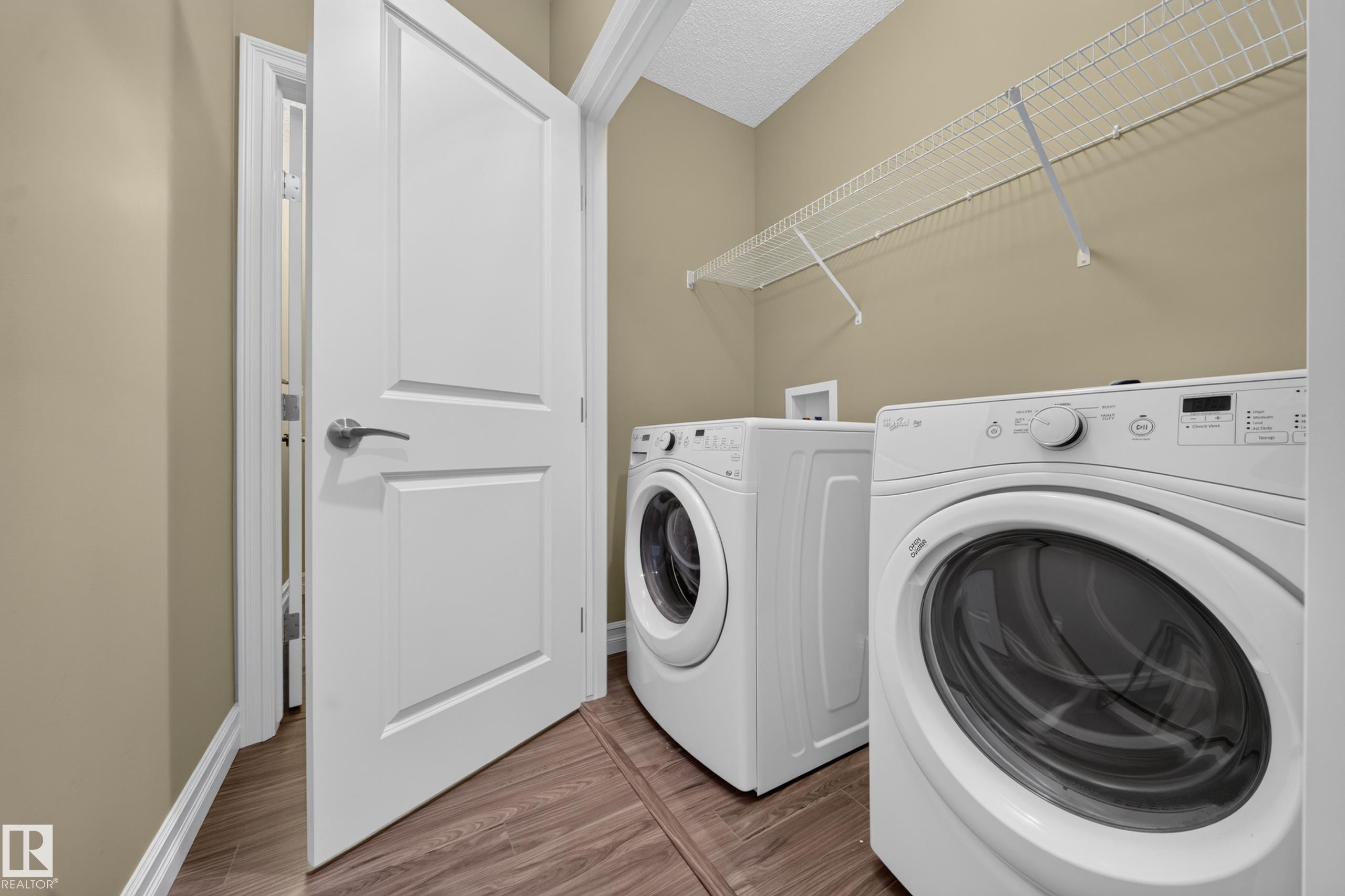 Dedicated laundry space featuring a front-load washer and dryer, wood-finish flooring, neutral-toned walls, and a wire storage shelf - #100 804 Welsh Dr Sw, Edmonton, AB - Indoor Photo Showing Laundry Room