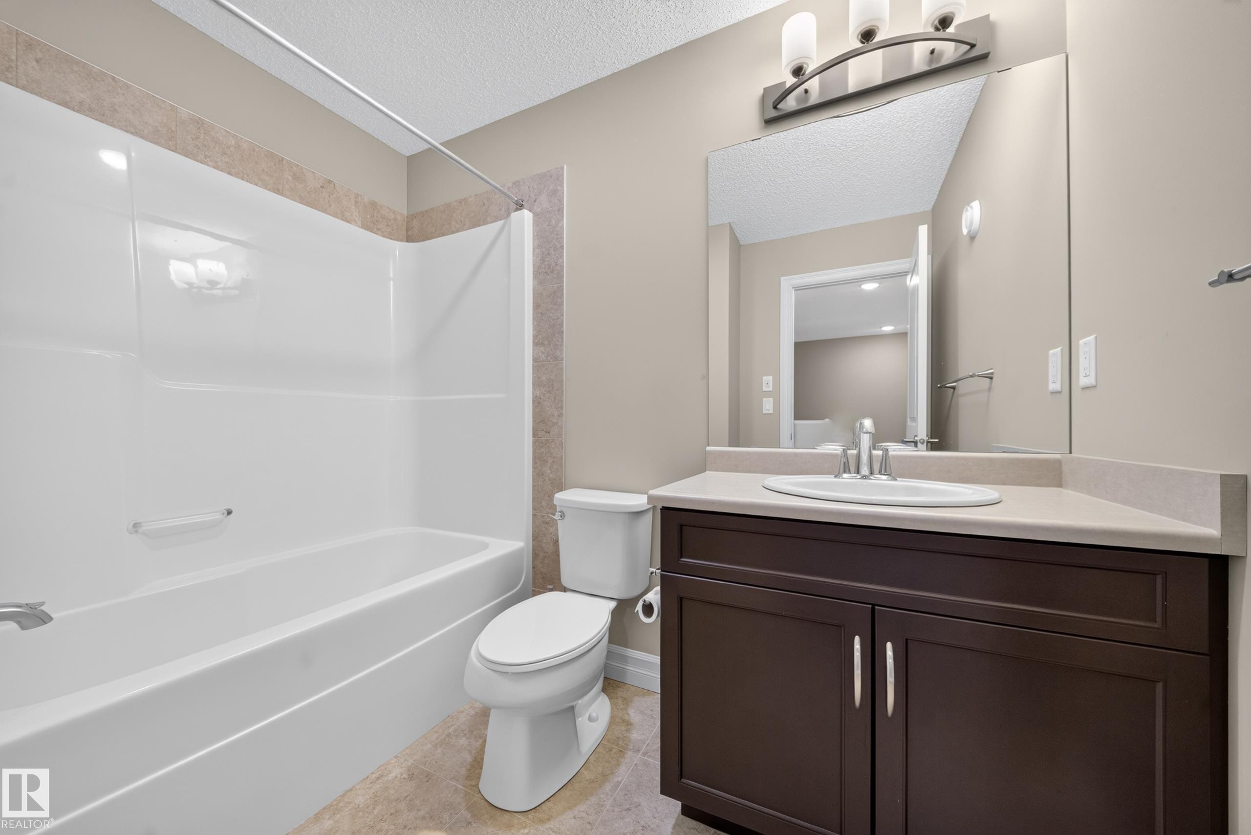 Bathroom featuring a white tub-shower combination with a tiled surround, a toilet, a dark wood-finish vanity with a light countertop, an undermount sink, and a large wall-mounted mirror - #100 804 Welsh Dr Sw, Edmonton, AB - Indoor Photo Showing Bathroom
