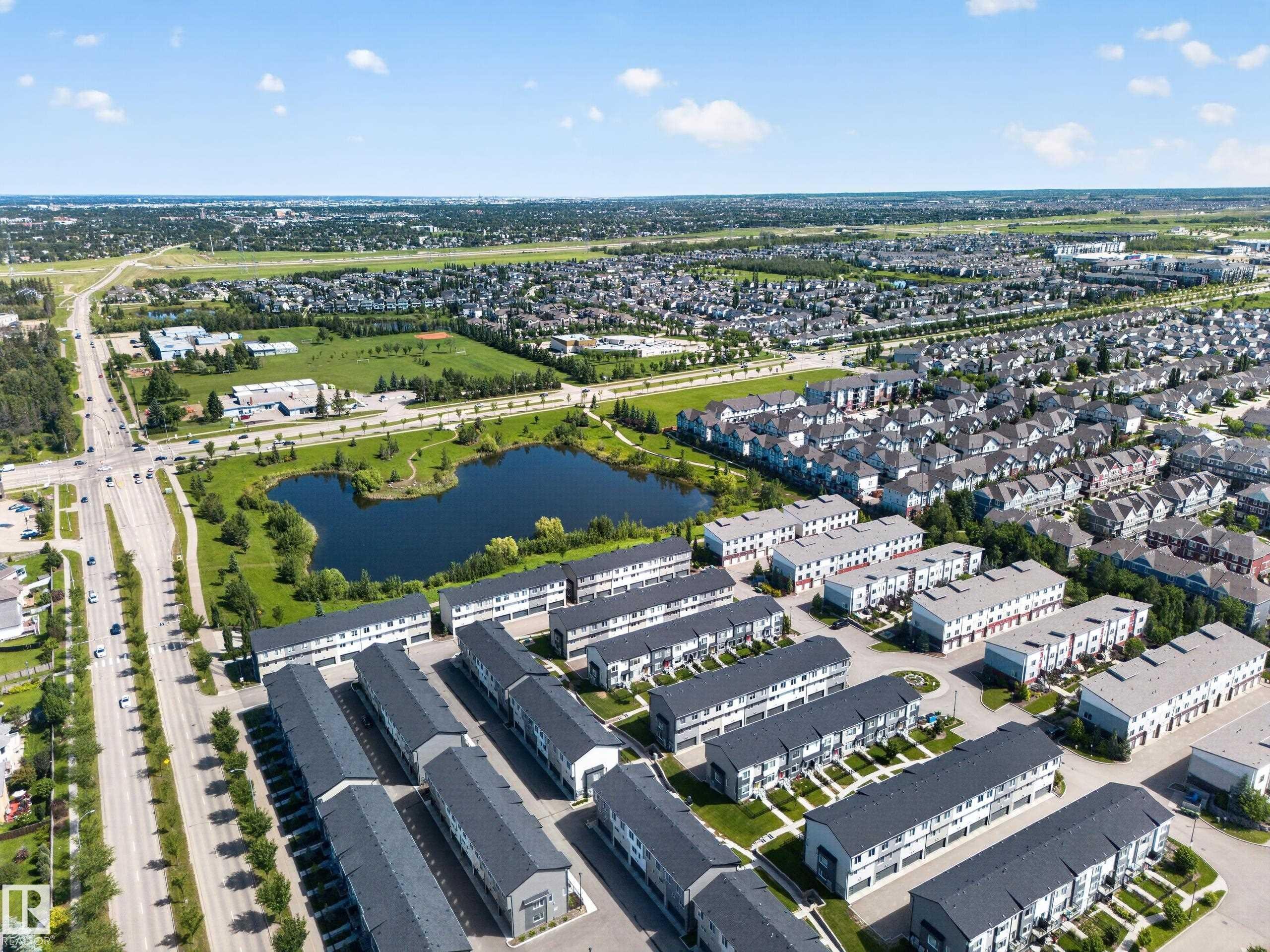 Aerial overview showcasing a developed community with modern townhomes, detached residences, and a central pond - #100 804 Welsh Dr Sw, Edmonton, AB - Outdoor With View