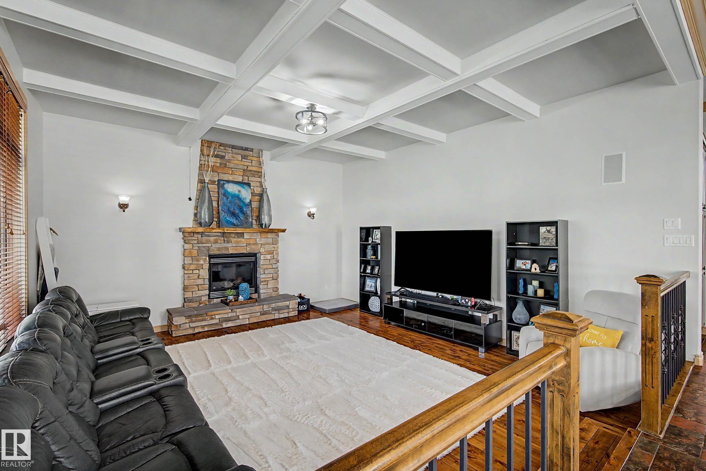Living area featuring a stone-clad fireplace, coffered ceiling, wood-finish flooring, and a wrought iron and wood staircase railing - 38 Longview Point(E), Spruce Grove, AB - Indoor Photo Showing Living Room With Fireplace