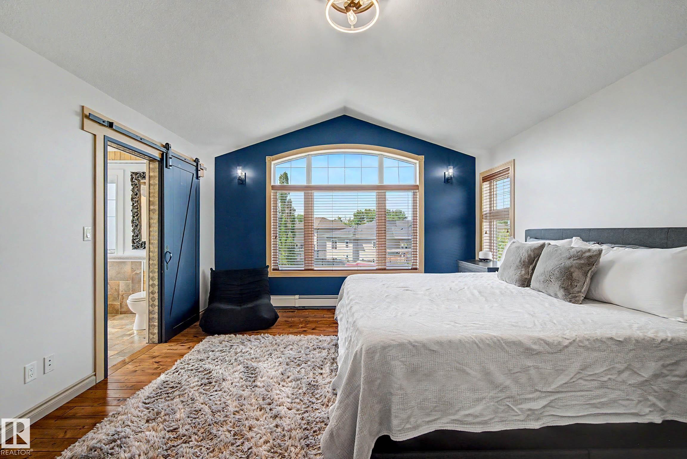 Vaulted ceiling bedroom featuring an arched window, wood-finish flooring, and a modern barn door leading to an adjacent bathroom - 38 Longview Point(E), Spruce Grove, AB - Indoor Photo Showing Bedroom