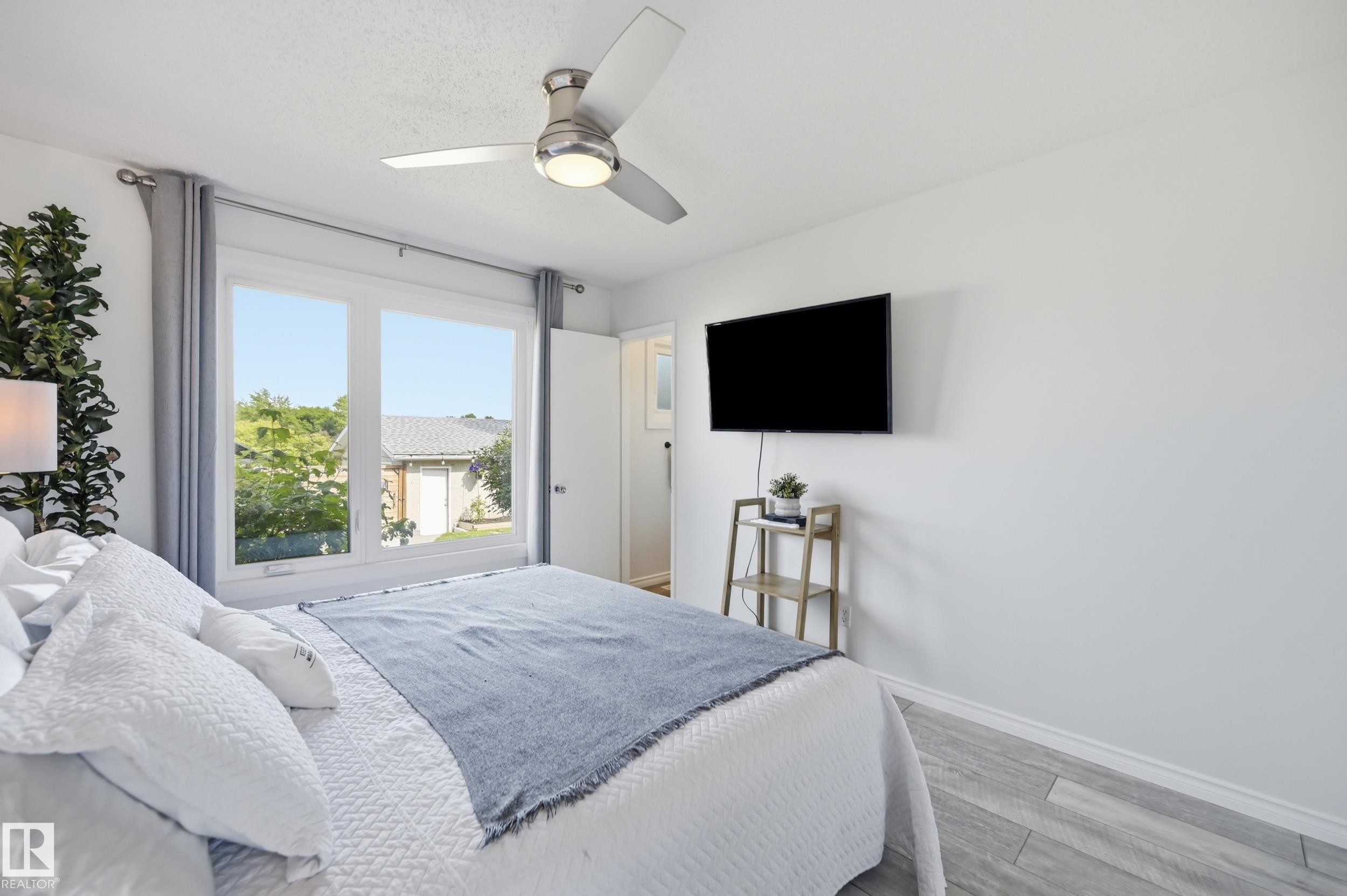 Bright interior with a large double-pane window, flush-mount ceiling fan, wall-mounted television, and wood-finish flooring - 15111 67 Street Nw, Edmonton, AB - Indoor Photo Showing Bedroom