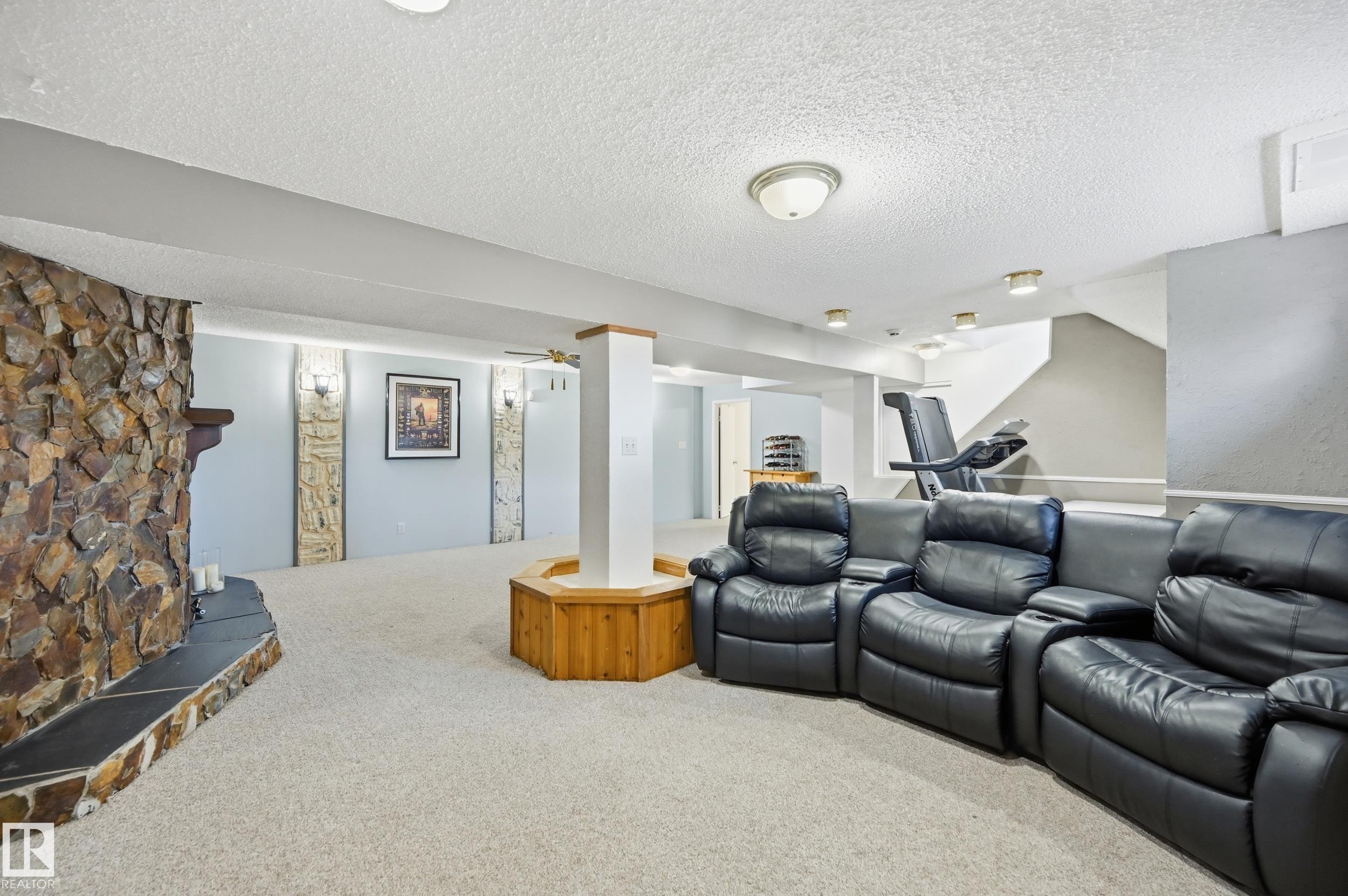 Expansive room featuring a stone-clad fireplace with a hearth, light-toned carpet, recessed lighting, ceiling-mounted light fixtures, and a staircase - 15111 67 Street Nw, Edmonton, AB - Indoor