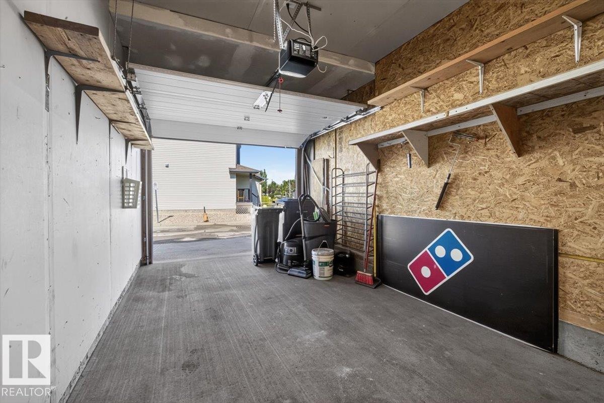 Garage interior with concrete flooring, overhead garage door with automatic opener, wood-finish shelving, and oriented strand board wall finish - #18 345 Kirkness Rd Nw, Edmonton, AB - Indoor Photo Showing Garage