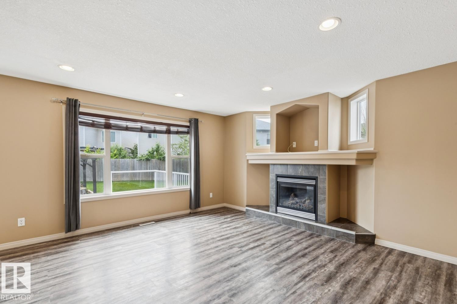 Spacious living area featuring wood-finish flooring, a corner fireplace with a tiled surround, and multiple recessed ceiling lights - 16765 119 Street Nw, Edmonton, AB