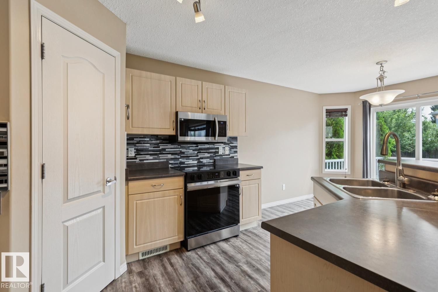Kitchen featuring light wood-finish cabinetry, stainless steel appliances, dark countertops, a mosaic tile backsplash, and wood-finish flooring - 16765 119 Street Nw, Edmonton, AB