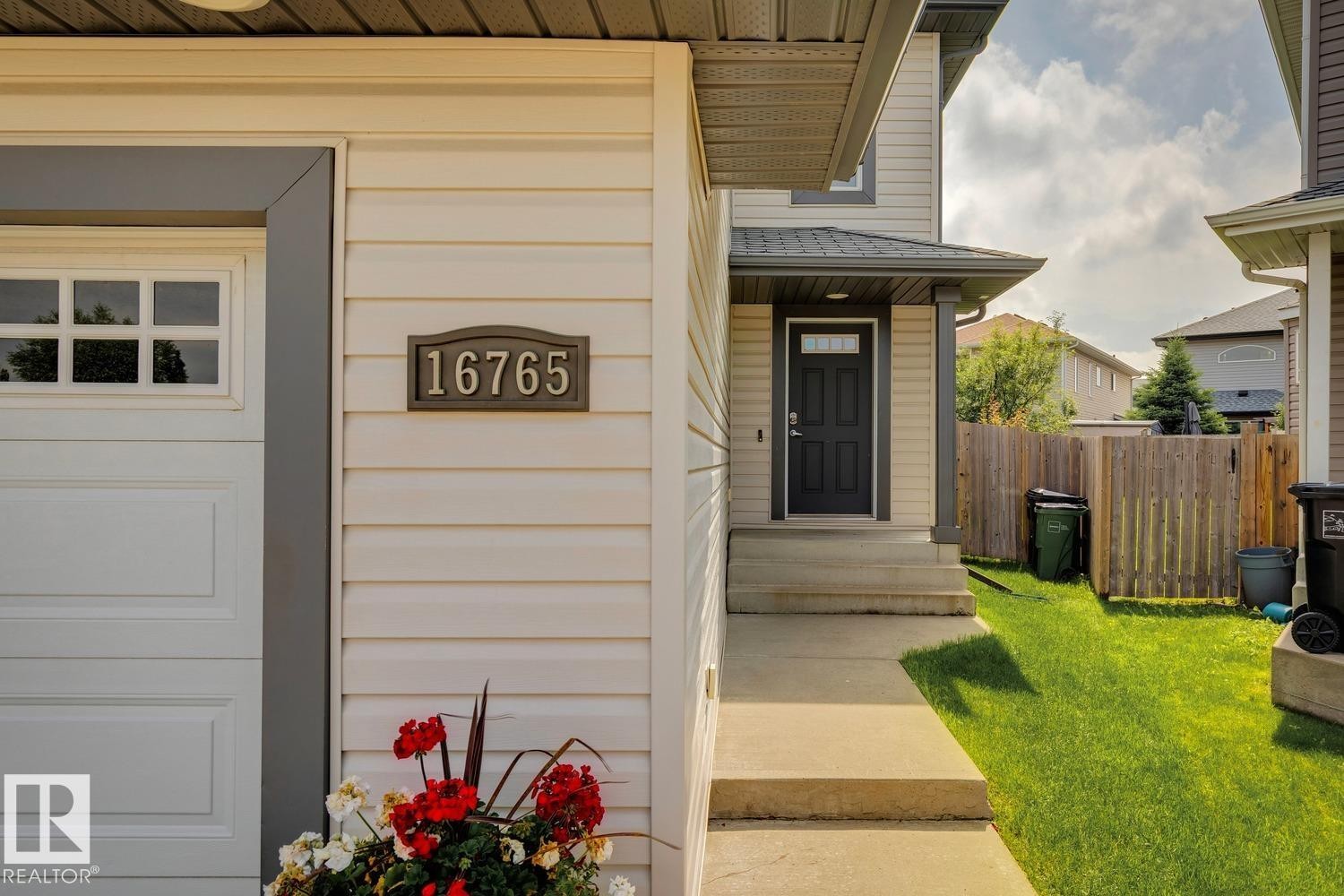 Exterior featuring light-toned siding, a dark front door with window inset, a white garage door with upper window paneling, a concrete walkway, and a partial grass lawn - 16765 119 Street Nw, Edmonton, AB