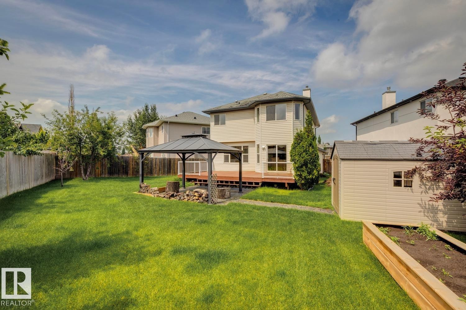 Expansive backyard featuring a large lawn, a wooden deck with a gazebo, a built-in fire pit area, a storage shed, and a raised garden bed - 16765 119 Street Nw, Edmonton, AB