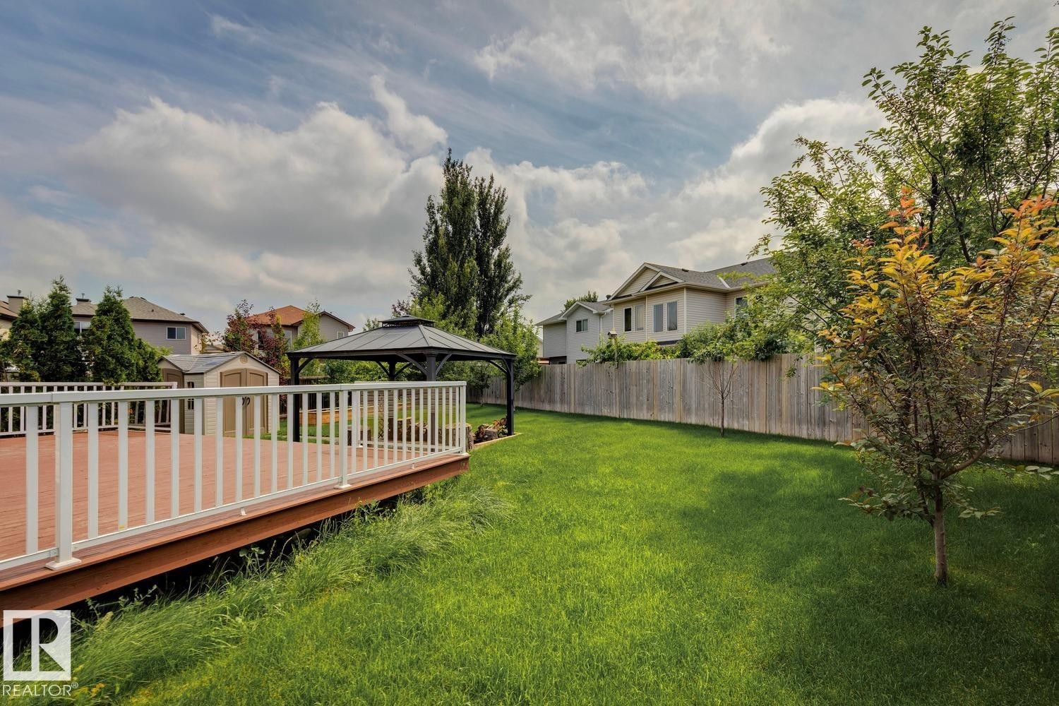 Expansive backyard featuring a wood-finish deck with white railings, a covered gazebo, a manicured lawn, and mature trees - 16765 119 Street Nw, Edmonton, AB