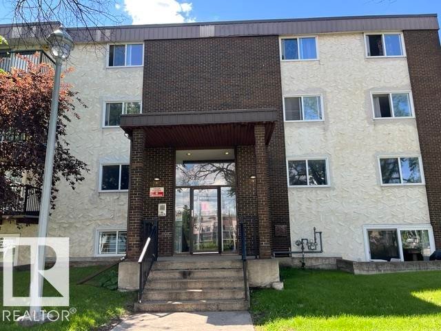 Building exterior featuring a brick facade with a stucco finish, multiple windows, a prominent entrance with steps, and surrounding green space - #205 10740 105 St Nw, Edmonton, AB - Outdoor