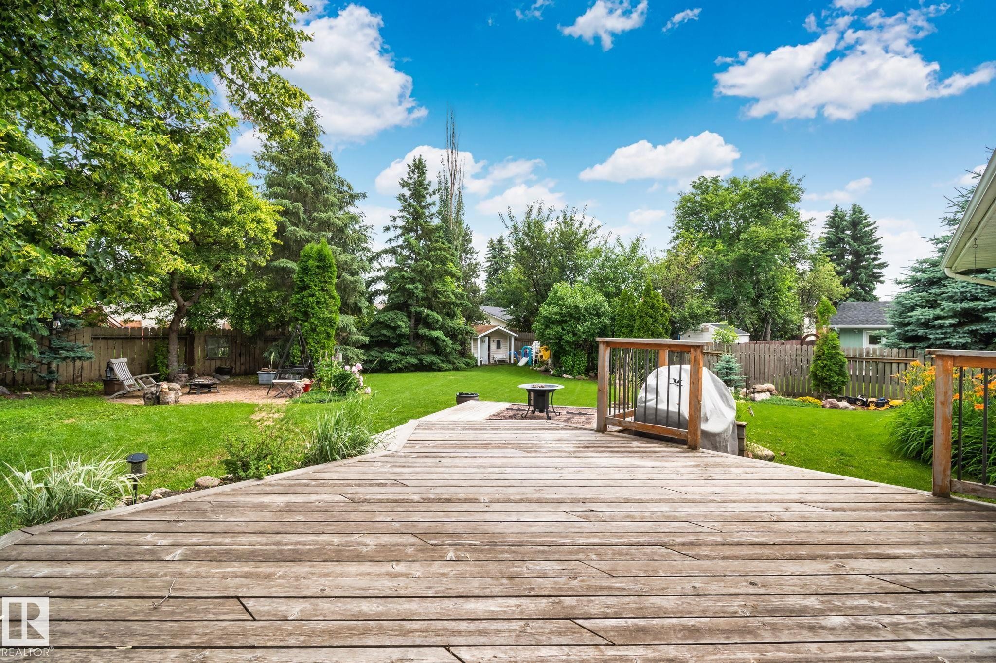 Spacious backyard featuring a large wooden deck with railings, an expansive lawn, mature trees, a fire pit area with stone paving, and a privacy fence - 7306 21 Avenue Nw, Edmonton, AB - Outdoor With Deck Patio Veranda