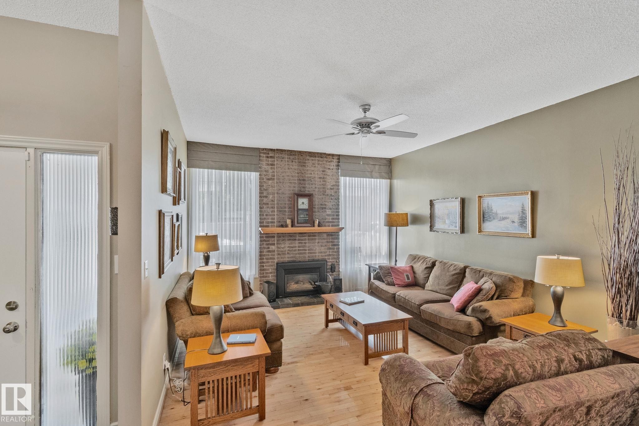Living area featuring a brick-surround fireplace with a wood mantle, wood-finish flooring, a ceiling fan, and a transom window - 7306 21 Avenue Nw, Edmonton, AB - Indoor Photo Showing Living Room With Fireplace