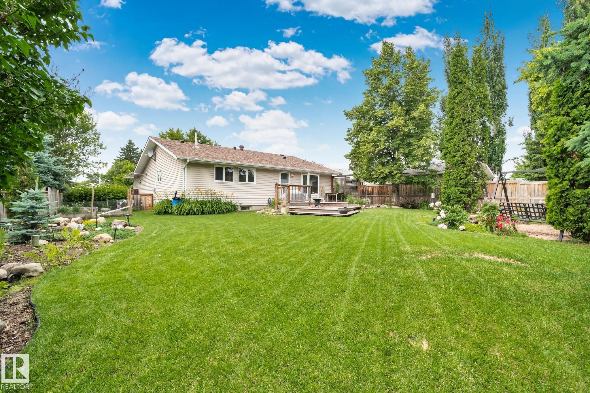 Spacious backyard featuring a large green lawn, a multi-level wood deck, mature perimeter trees, and a rock-lined garden bed - 7306 21 Avenue Nw, Edmonton, AB - Outdoor