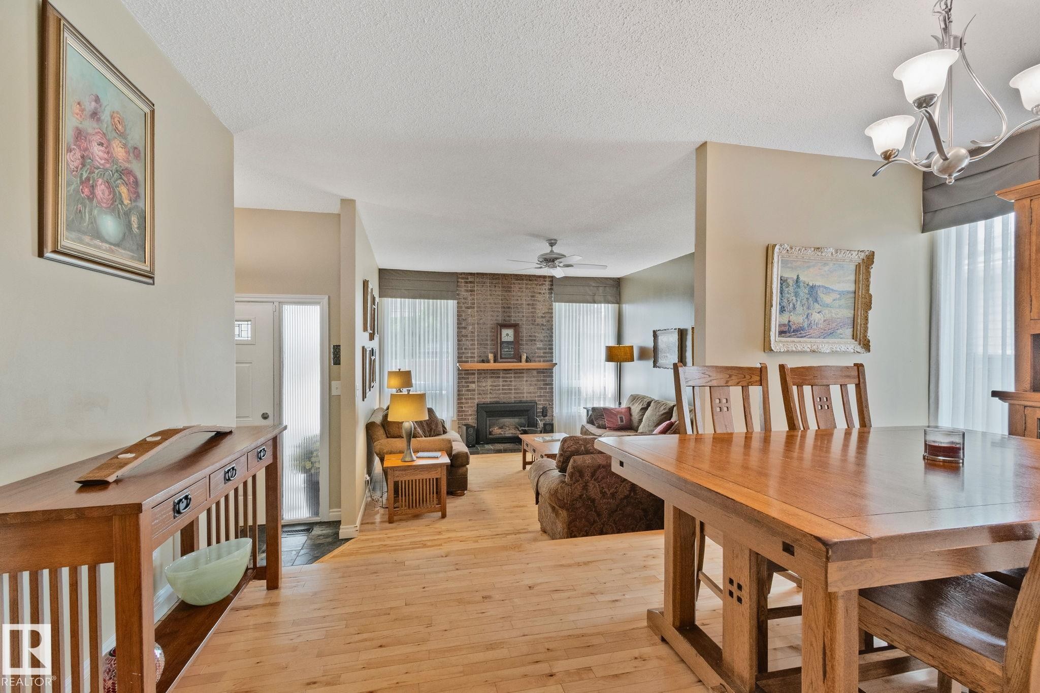 Open-concept interior featuring wood-finish flooring, a dining area with a wood table, and a living area with a brick fireplace - 7306 21 Avenue Nw, Edmonton, AB - Indoor Photo Showing Dining Room With Fireplace