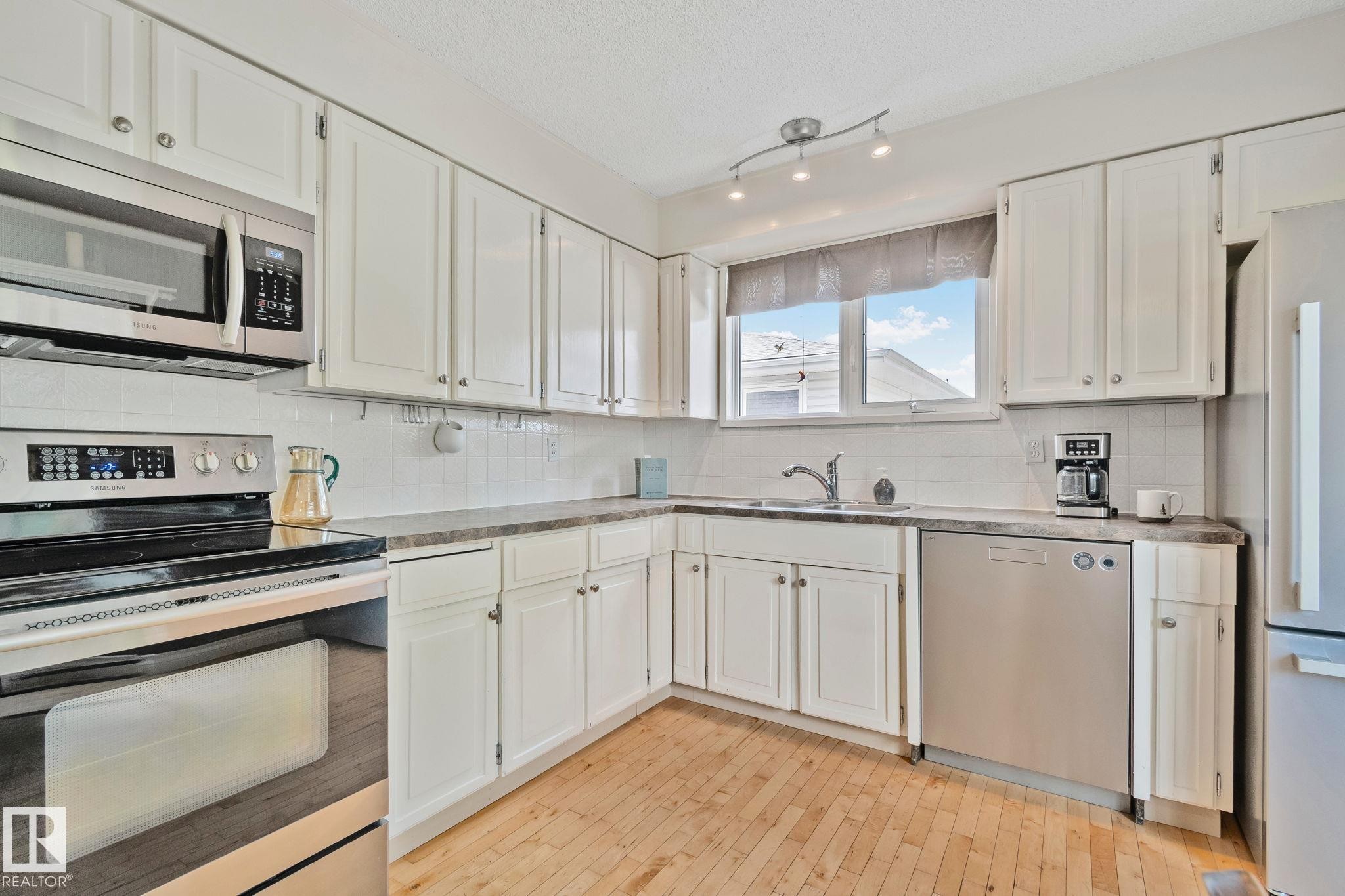 Kitchen featuring white cabinetry, stainless steel appliances, wood-finish flooring, a white subway tile backsplash, and a window with natural light - 7306 21 Avenue Nw, Edmonton, AB - Indoor Photo Showing Kitchen With Double Sink