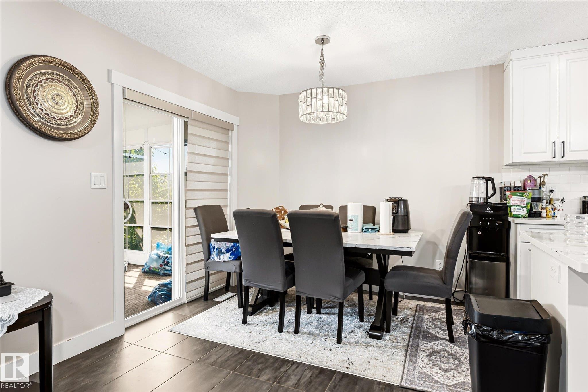 Dining area with dark wood-finish flooring, featuring a modern chandelier and direct access to the exterior through a glass sliding door with horizontal blinds - 4317 152 Avenue Nw, Edmonton, AB - Indoor Photo Showing Dining Room