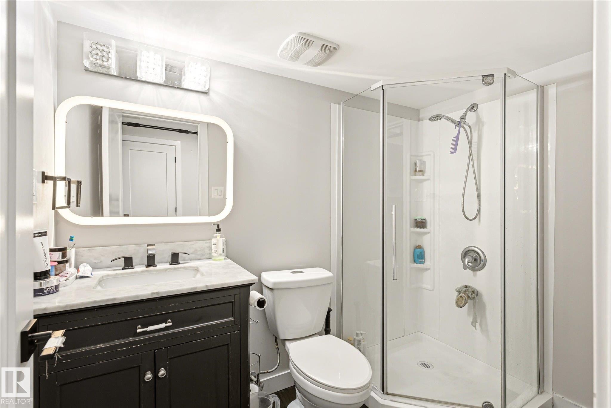 Bathroom featuring a corner glass-enclosed shower, a vanity with a white countertop and dark cabinetry, a wall-mounted mirror with integrated lighting, and a brushed nickel faucet - 4317 152 Avenue Nw, Edmonton, AB - Indoor Photo Showing Bathroom
