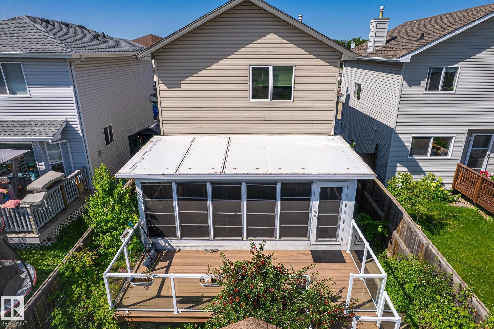 Rear exterior featuring a sunroom with a white framed door, a wood-finish deck with glass panel railing, and mature green landscaping - 4317 152 Avenue Nw, Edmonton, AB - Outdoor With Exterior
