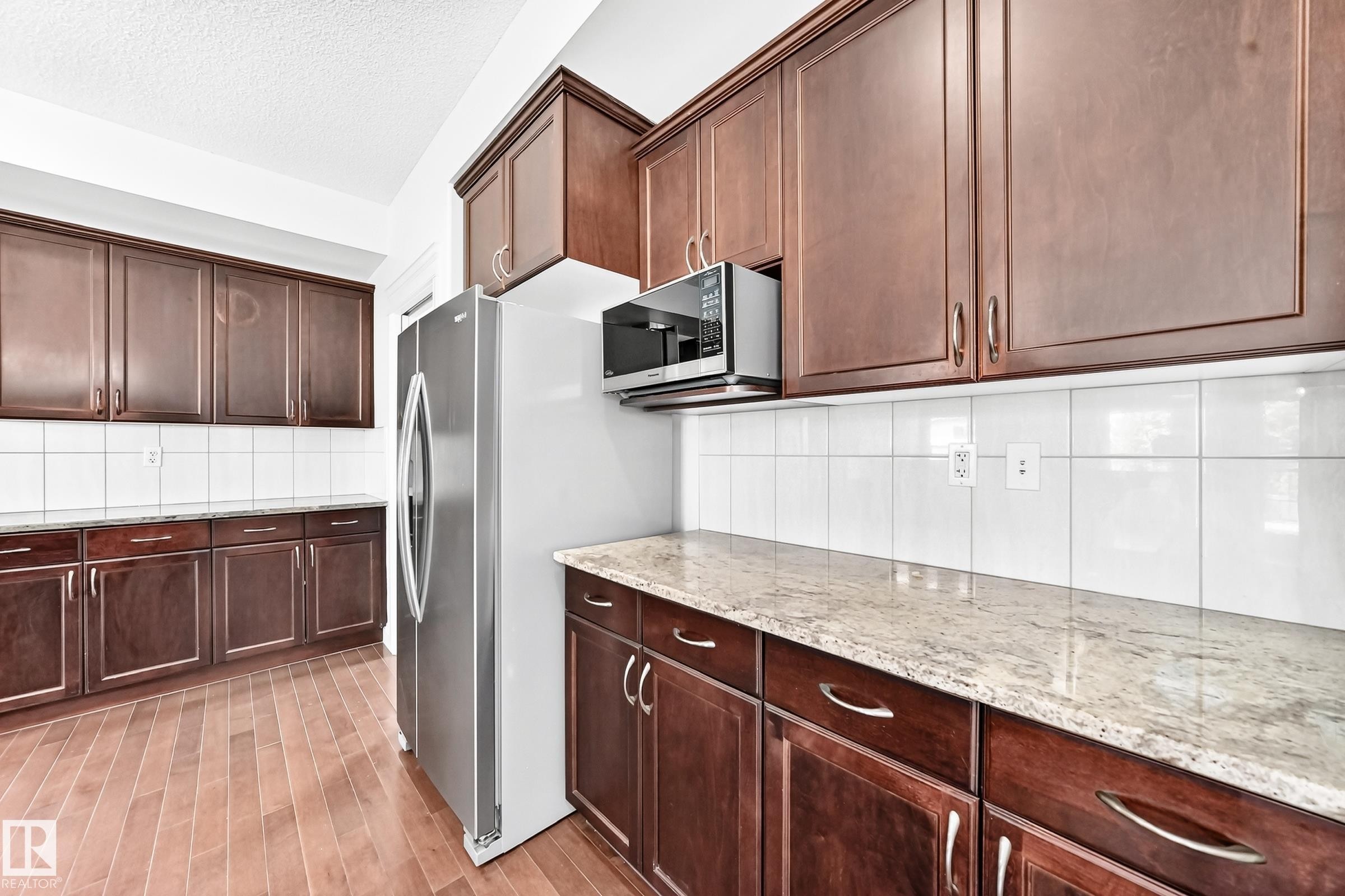 Kitchen featuring wood-finish flooring, dark wood cabinetry, light-colored countertops, a white tile backsplash, and a stainless steel refrigerator - 1663 Rutherford Road Sw, Edmonton, AB - Indoor Photo Showing Kitchen