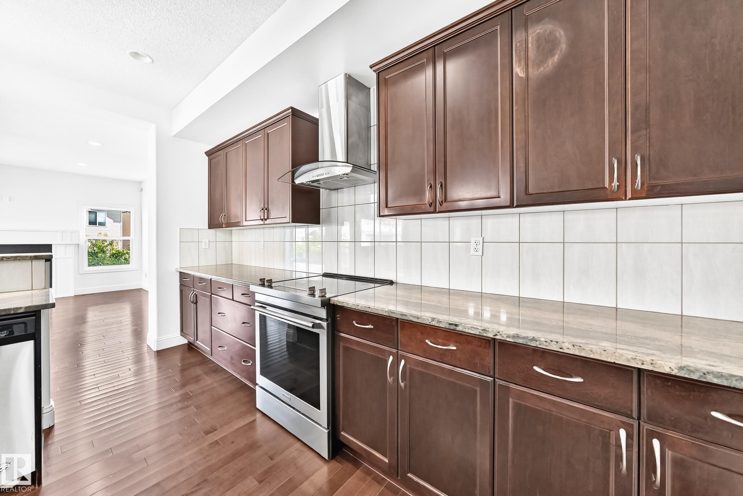 Kitchen featuring wood-finish flooring, dark wood cabinetry, light-toned countertops, a tile backsplash, and stainless steel appliances - 1663 Rutherford Road Sw, Edmonton, AB - Indoor Photo Showing Kitchen