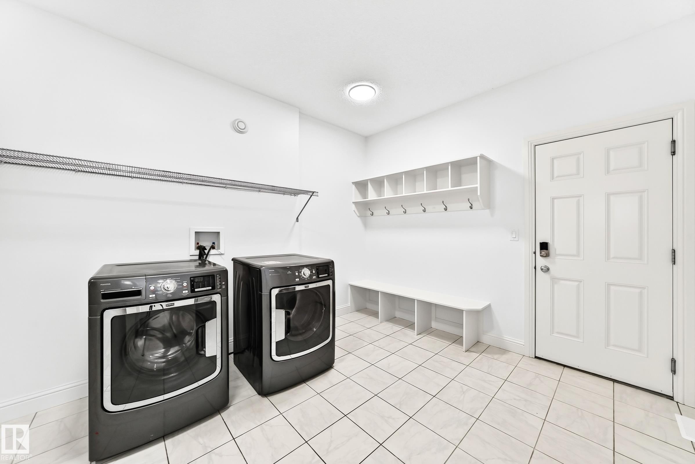 Utility room featuring a white paneled door, built-in shelving with coat hooks, a built-in bench, and light-toned tile flooring - 1663 Rutherford Road Sw, Edmonton, AB - Indoor Photo Showing Laundry Room