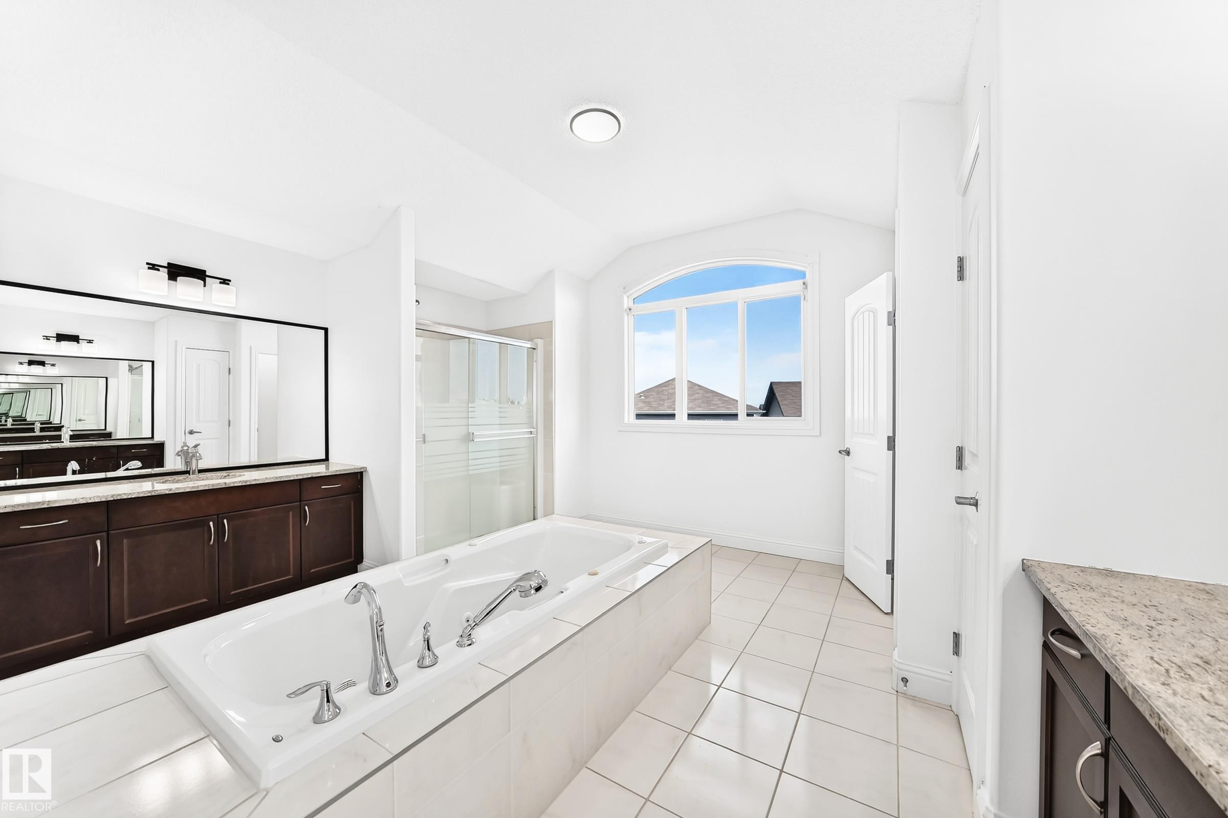 Spacious bathroom featuring a built-in soaking tub, separate glass-enclosed shower, dual vanities with dark wood cabinetry, and an arched window - 1663 Rutherford Road Sw, Edmonton, AB - Indoor