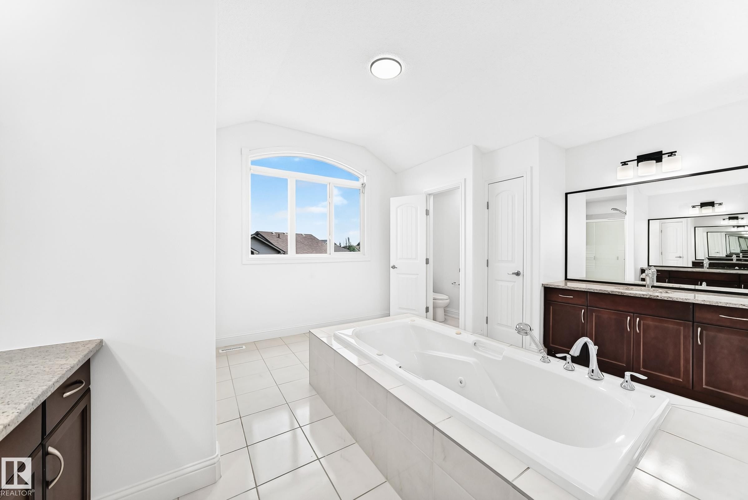 Spacious bathroom featuring a built-in soaking tub with chrome fixtures, dual vanity with dark wood cabinetry and light-toned countertops, and an arched window - 1663 Rutherford Road Sw, Edmonton, AB - Indoor Photo Showing Bathroom