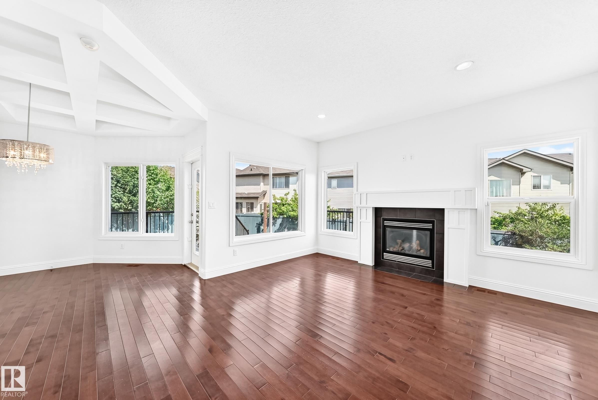 Bright living area featuring wood-finish flooring, multiple large windows, a central fireplace with a dark surround and white mantel, a decorative coffered ceiling detail, and a contemporary chandelier - 1663 Rutherford Road Sw, Edmonton, AB - Indoor Photo Showing Living Room With Fireplace