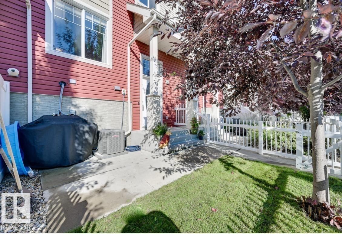 Exterior featuring red horizontal siding, a concrete patio, a manicured lawn, a mature tree, and a white picket fence - #16 460 Hemingway Rd Nw, Edmonton, AB - Outdoor