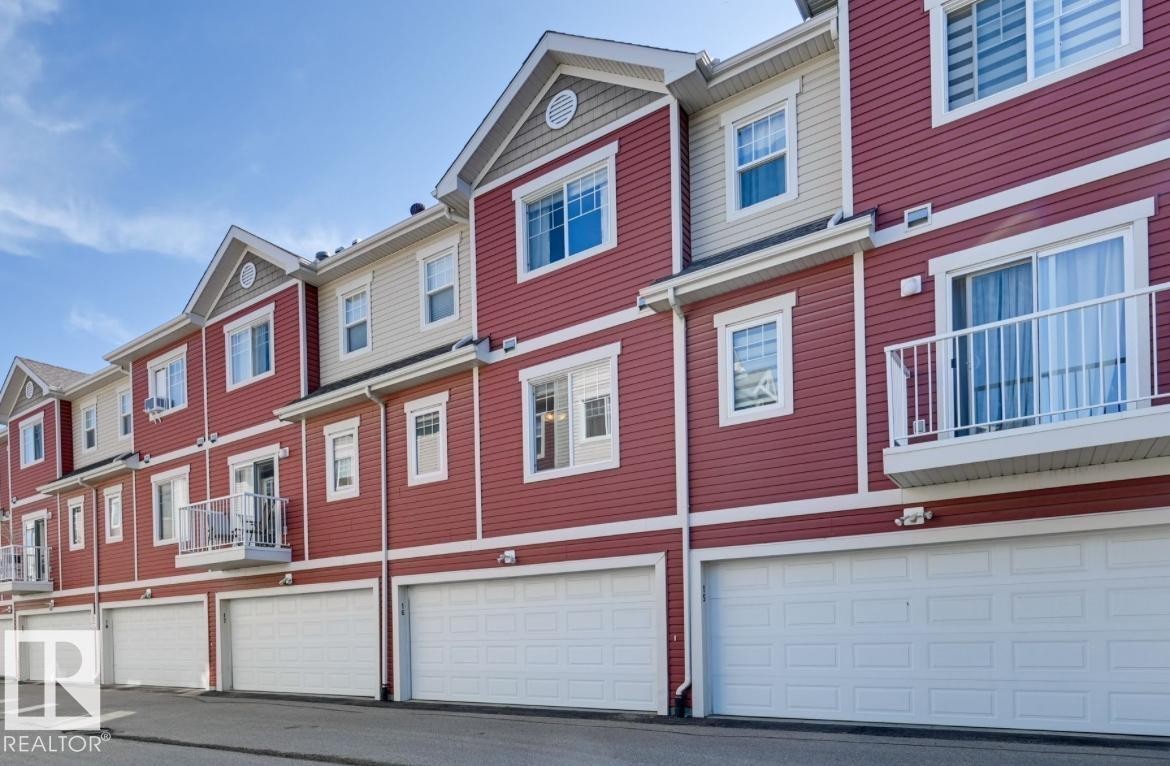 Townhome exterior featuring red and beige siding, individual garage doors, multiple windows, and a private balcony with white railings - #16 460 Hemingway Rd Nw, Edmonton, AB - Outdoor