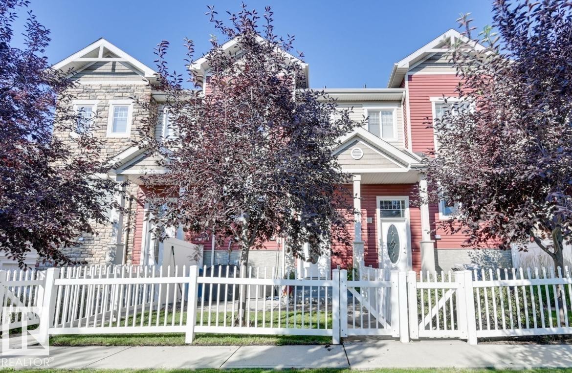 Multi-level facade featuring stone and siding finishes, a covered entryway with columns, a white picket fence, and mature trees providing shade - #16 460 Hemingway Rd Nw, Edmonton, AB - Outdoor