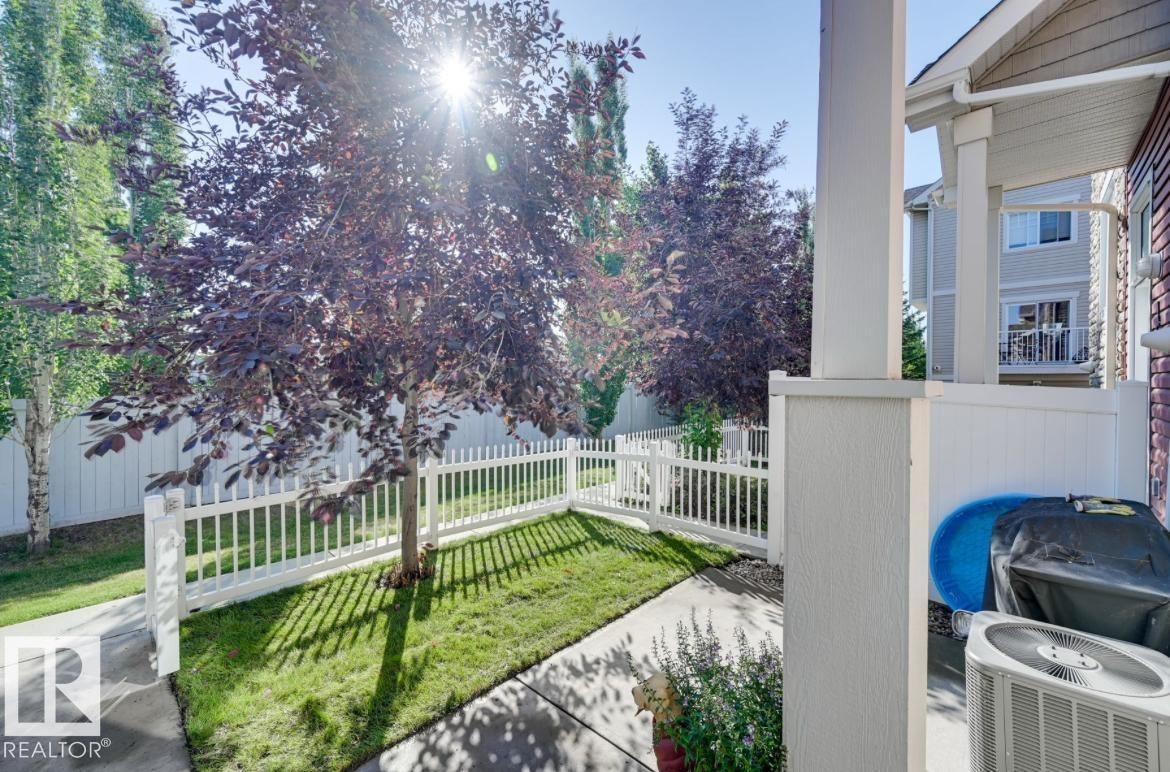 Landscaped side yard featuring mature trees, a white picket fence, and concrete paving - #16 460 Hemingway Rd Nw, Edmonton, AB - Outdoor