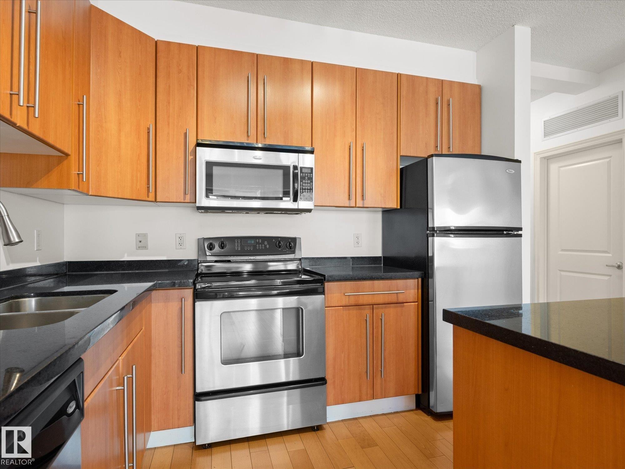 Kitchen featuring wood-finish cabinetry, dark countertops, and stainless steel appliances including a refrigerator, range, microwave, and dishwasher - #707 9819 104 St Nw, Edmonton, AB