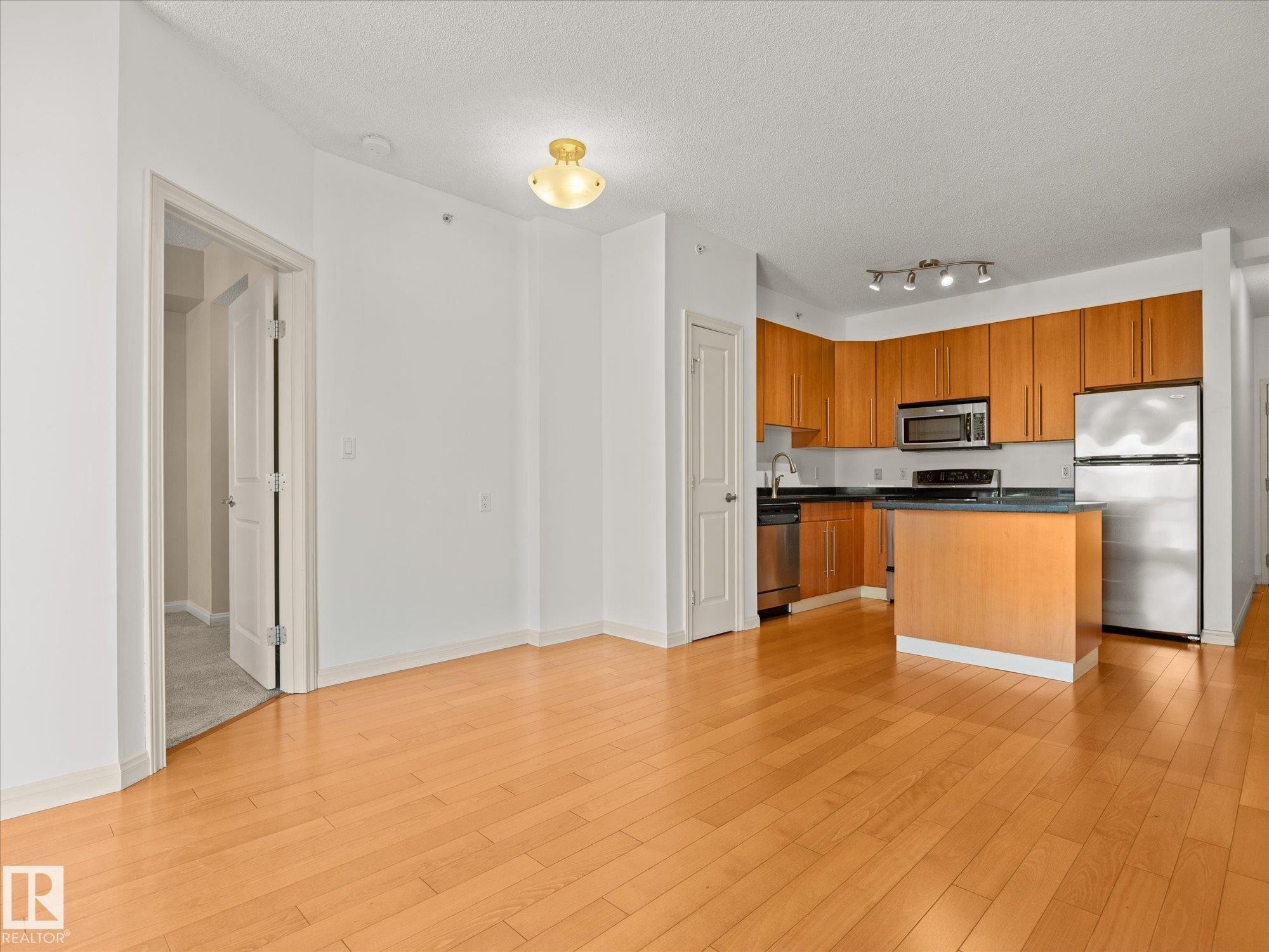 Open-concept kitchen featuring wood-finish cabinetry, stainless steel appliances, dark countertops, and a central island - #707 9819 104 St Nw, Edmonton, AB