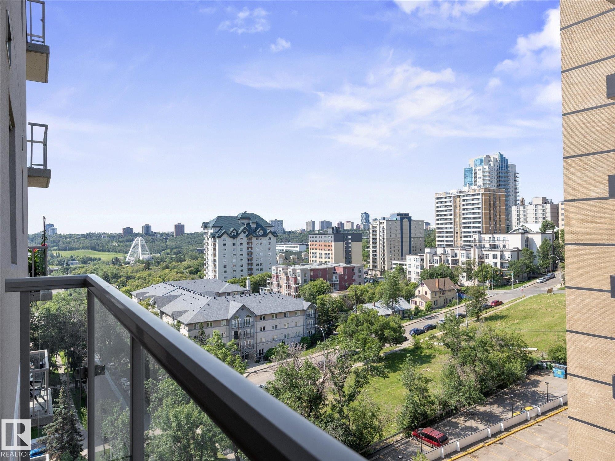Glass-paneled balcony railing providing an expansive urban landscape with distant architectural structures and lush green spaces - #707 9819 104 St Nw, Edmonton, AB